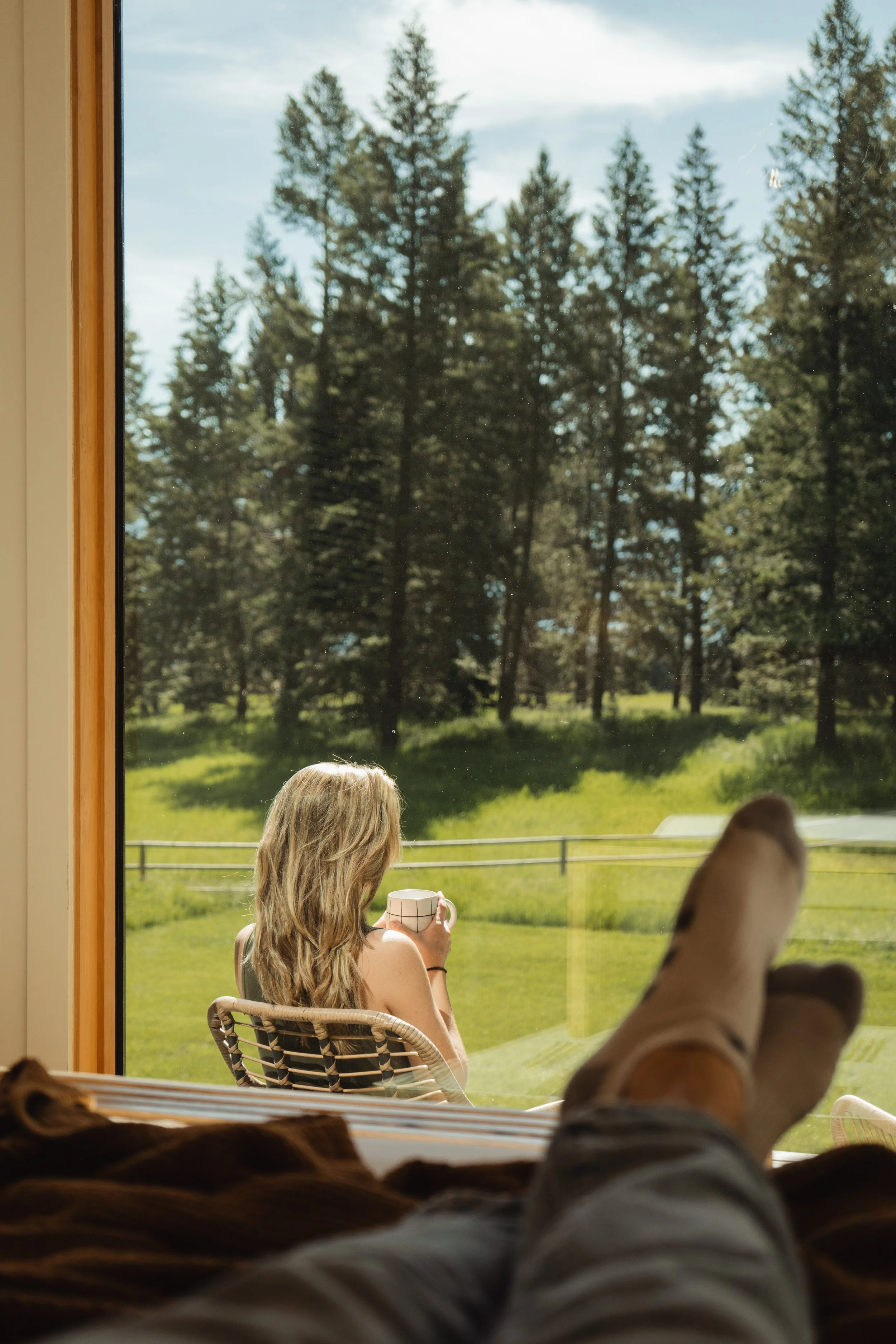 View from inside a house showing a woman sitting outside on a chair, holding a mug, with a window frame and a scenic landscape of green grass, trees, and a fence outside.