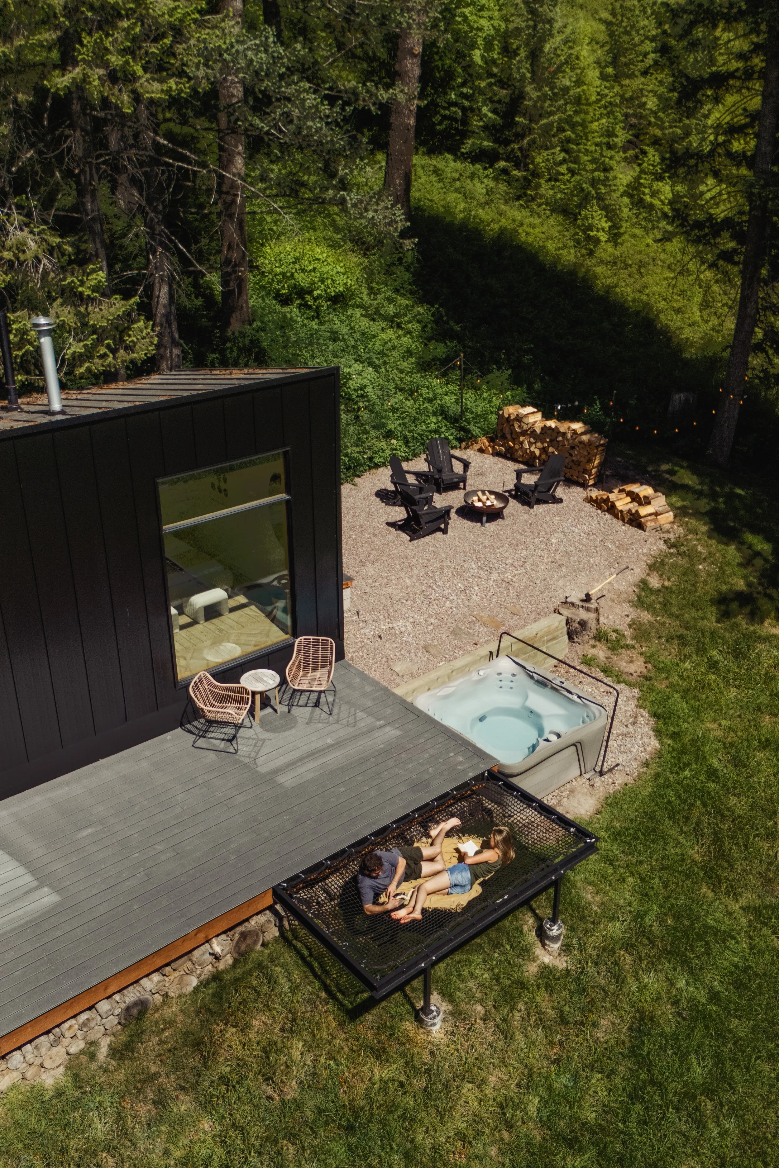 A backyard with a deck attached to a modern black house, a hot tub, a lounge area with two people laying on a outdoor lounge, and a gravel yard with stacked firewood, surrounded by tall green trees.
