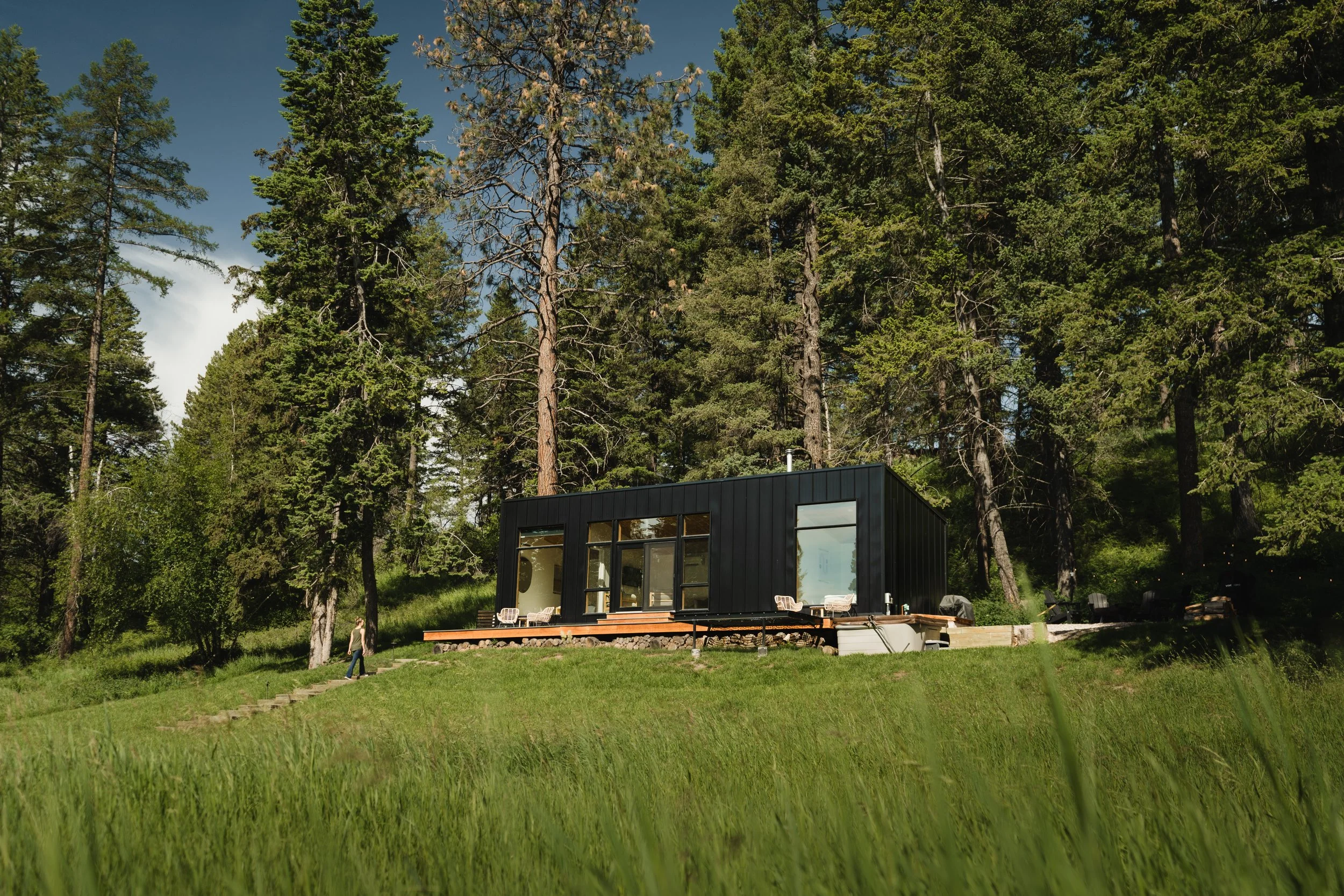 Modern black house with large windows, surrounded by tall pine trees and green grass, with outdoor seating on a deck.