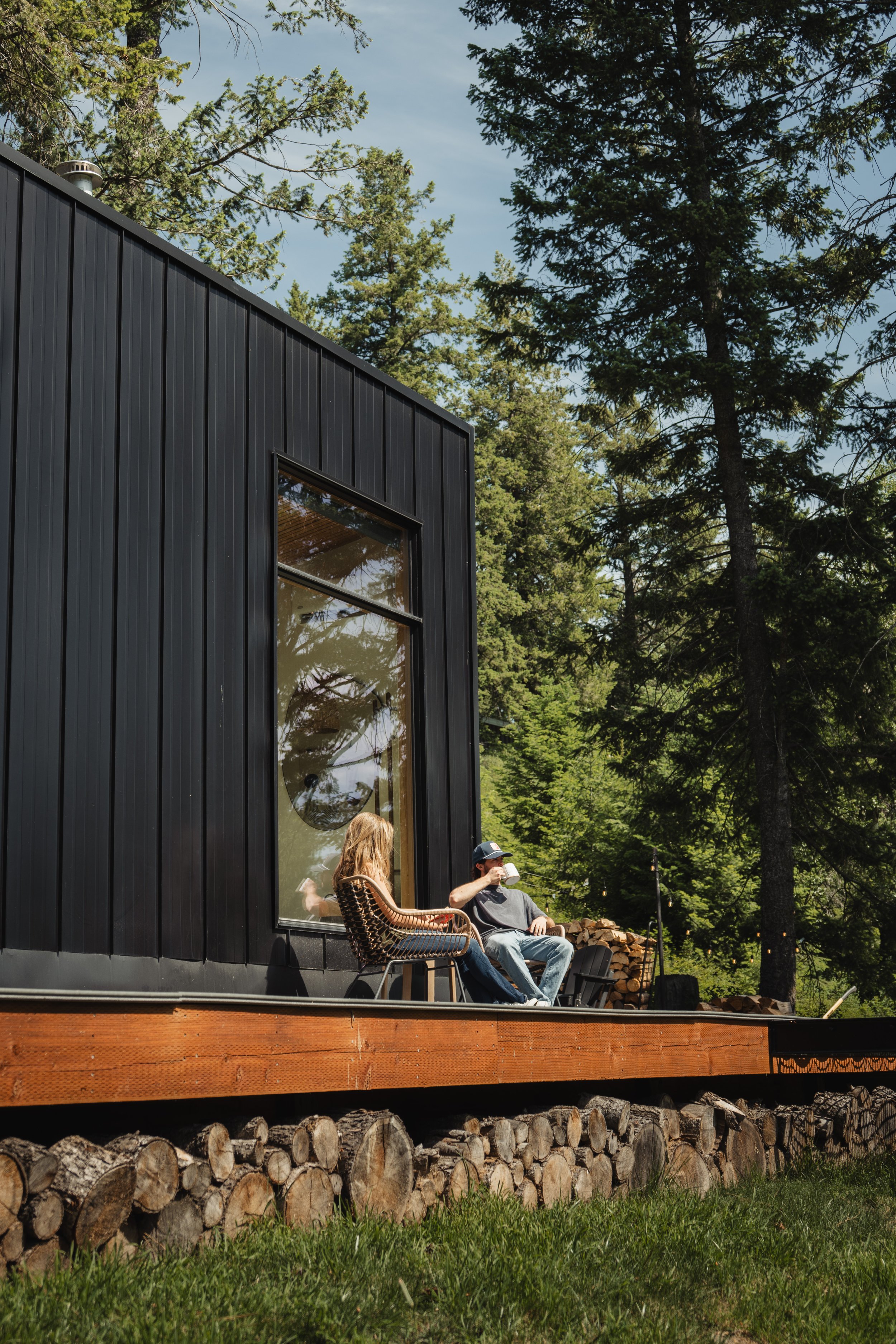 Two people sitting outside on a wooden deck, talking and drinking coffee or tea, with a black modern house and tall green trees in the background.