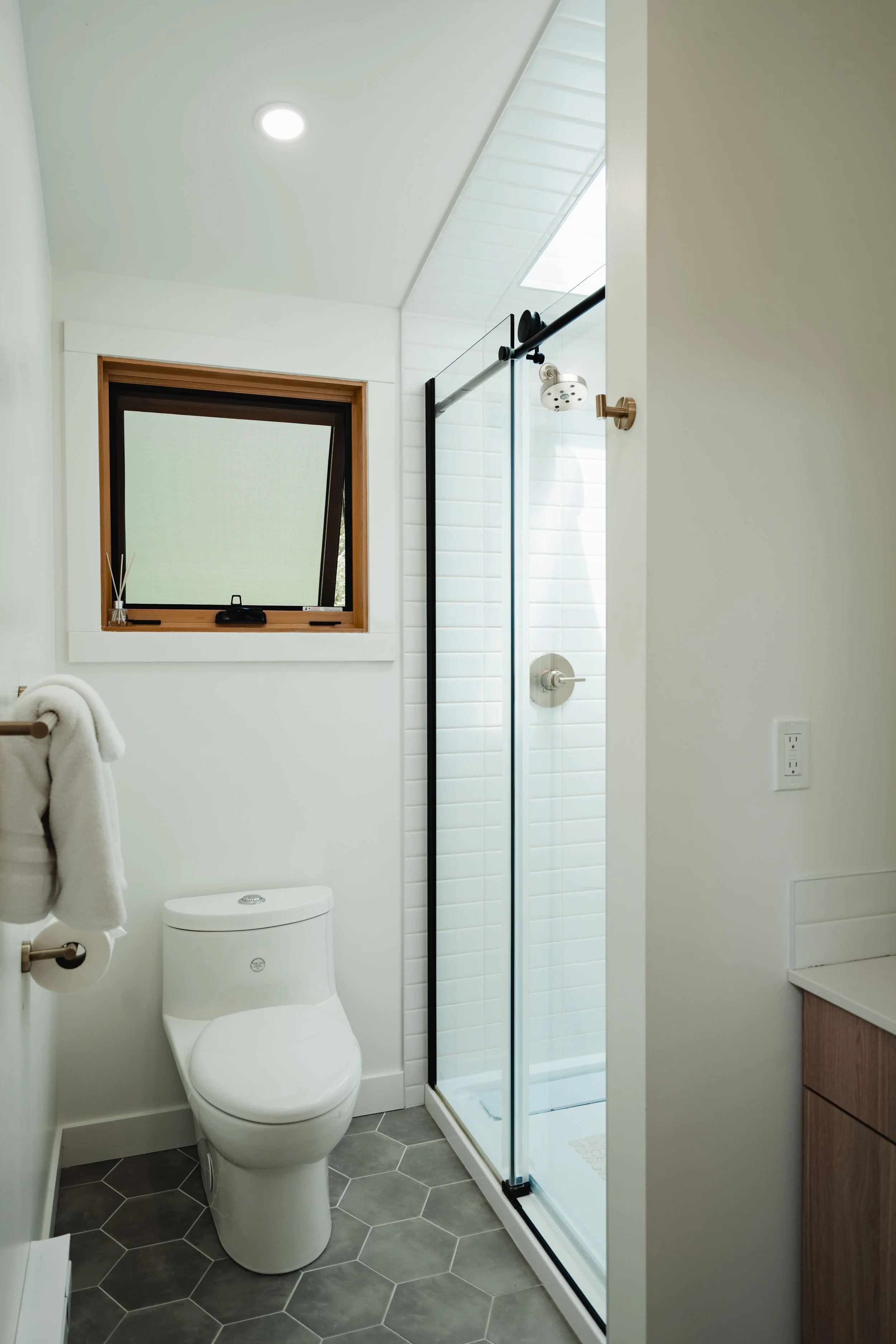 A modern bathroom with a white toilet, a small window, a glass-enclosed shower, gray hexagonal floor tiles, and a wooden cabinet.