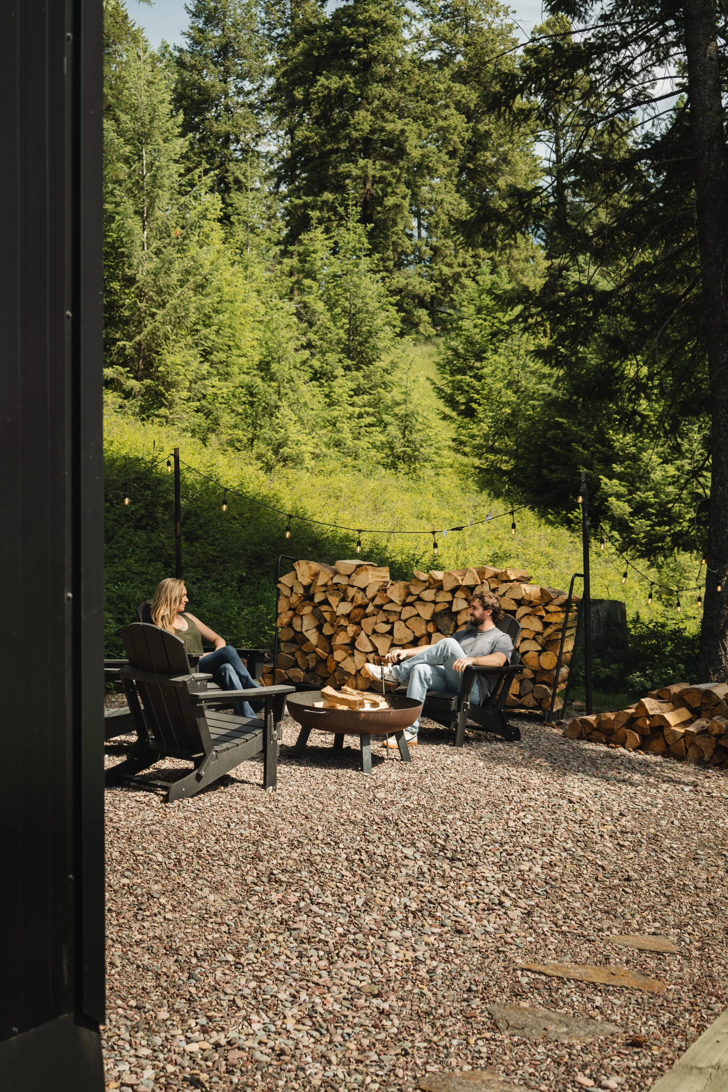 Two people sitting in outdoor chairs, talking, with a large stack of firewood and trees in the background, surrounded by string lights, on a gravel ground.