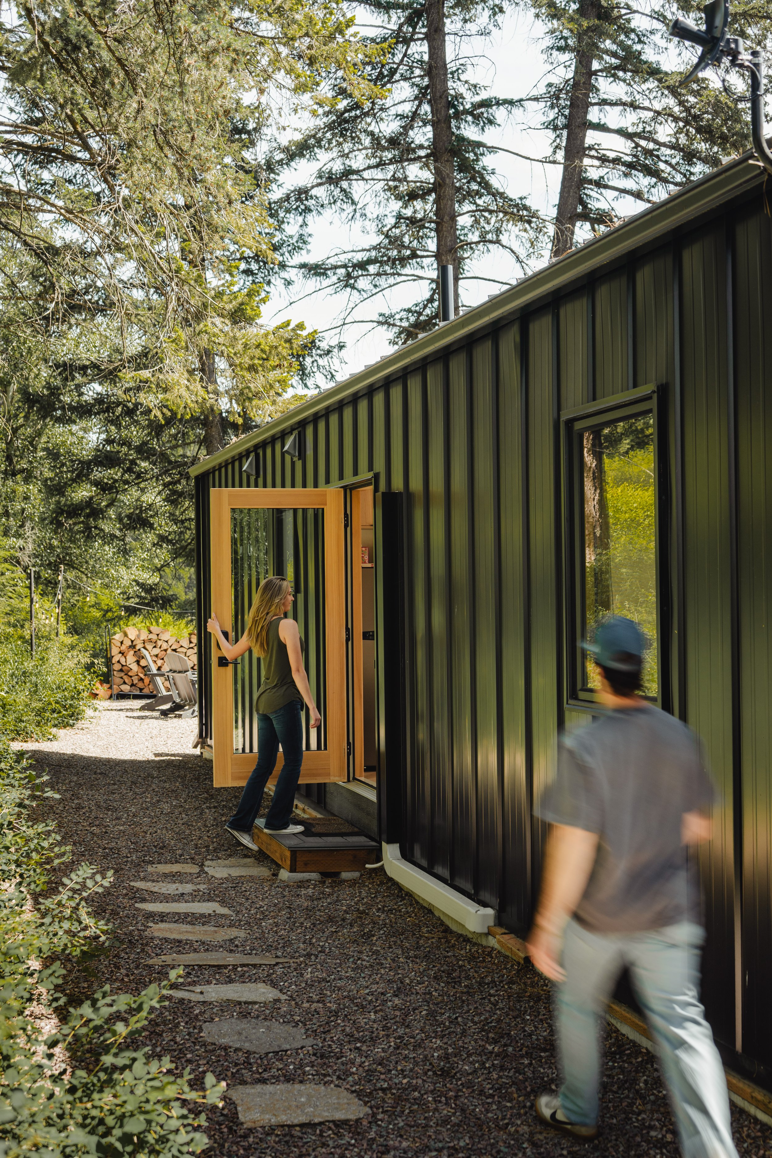 A woman is standing on a porch next to a modern black metal house, opening the door. Two blurred people are walking nearby in a garden with trees, shrubs, and a stone pathway.