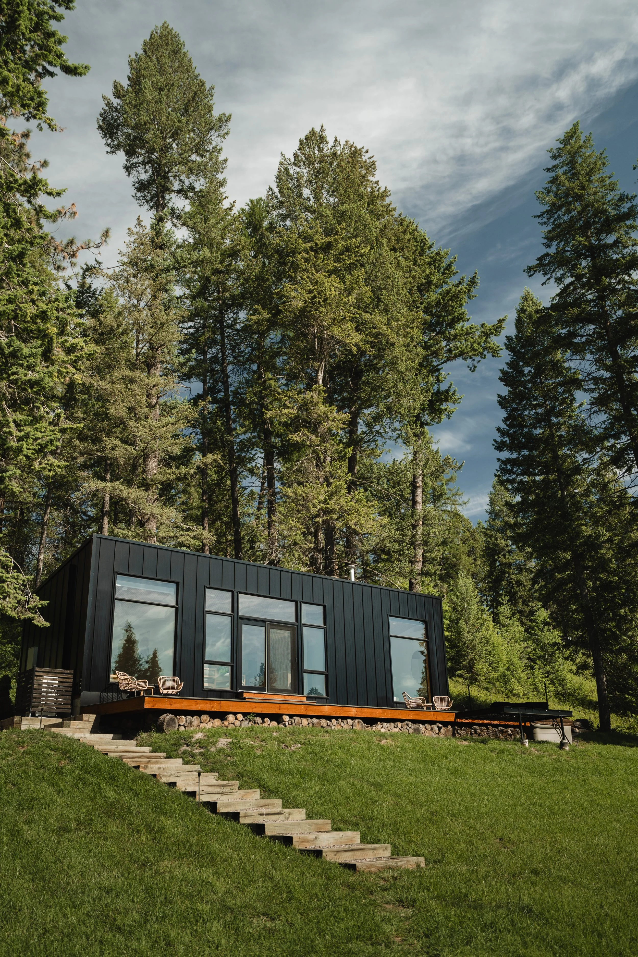 Modern black house with large windows, surrounded by lush green grass and trees, with a hilly forested background under a partly cloudy sky.
