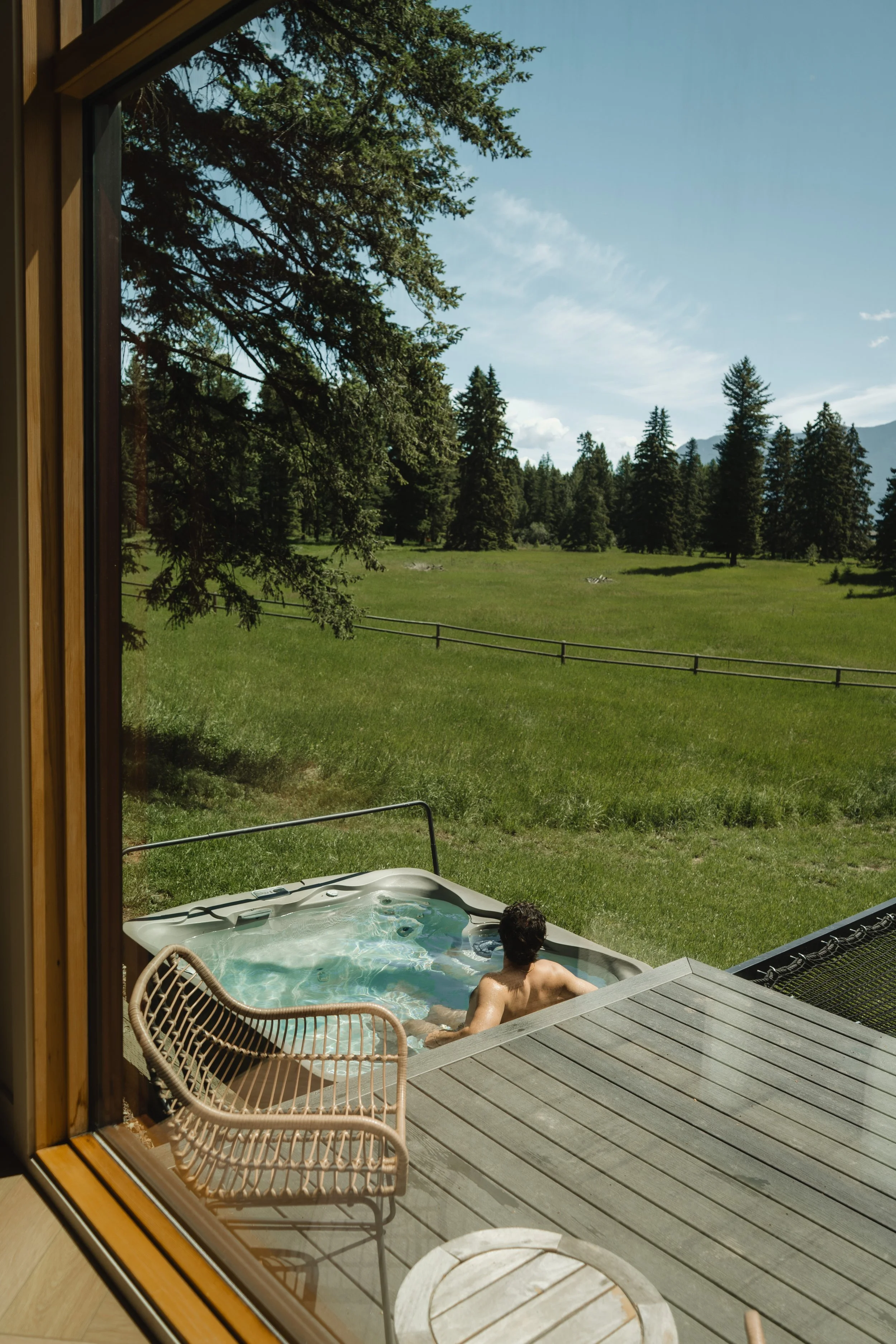 A man is relaxing in a hot tub on a wooden deck, looking out at a lush green field with tall trees and mountains in the distance on a sunny day.