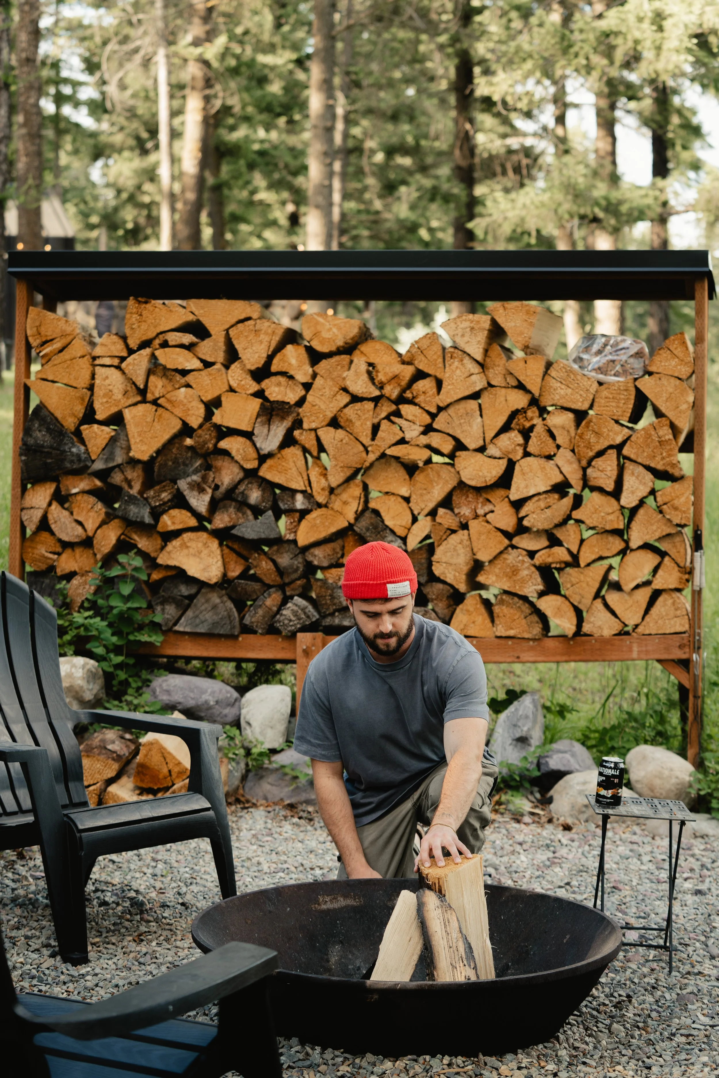 A man with a beard and a red beanie is kneeling and chopping a piece of wood in a fire pit outdoors, with stacked firewood and forest trees in the background.