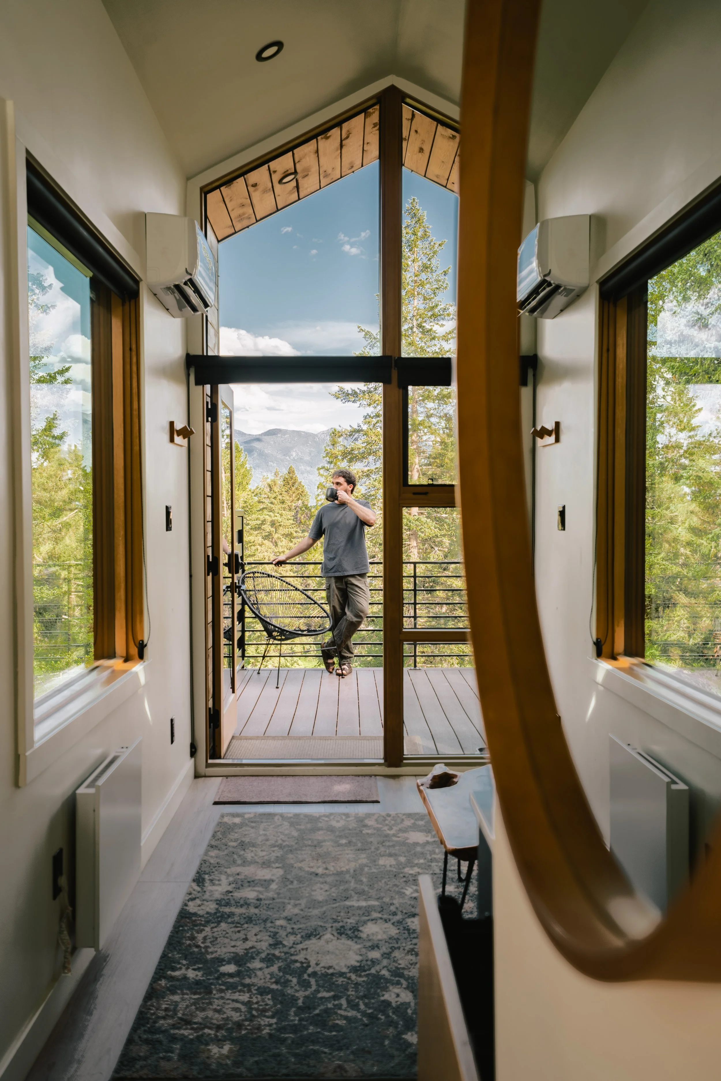 A person standing on a balcony, drinking coffee, with mountains and trees visible in the background through open glass doors and windows.