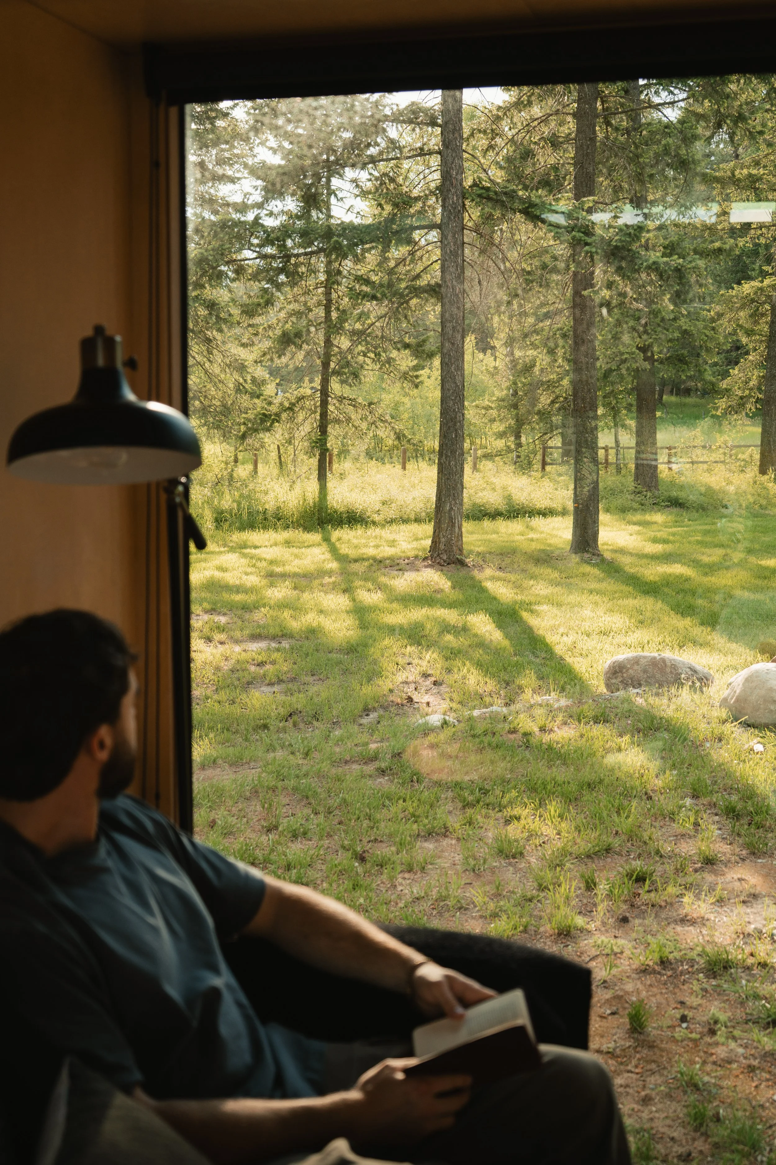 A man sitting indoors near a large window, reading a book, with a view of trees and grass outside during the daytime.
