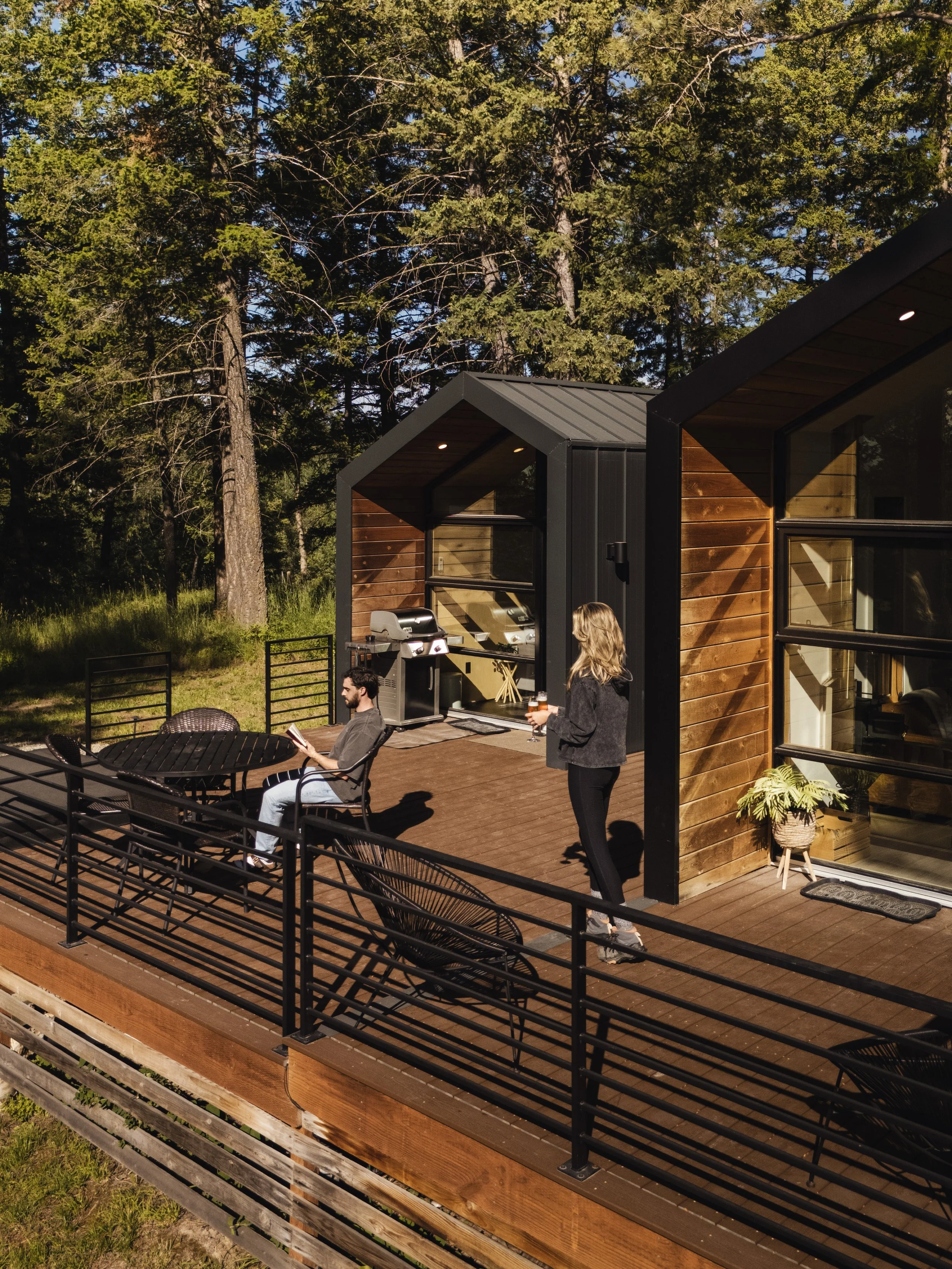 A modern house with large windows and a wooden deck, surrounded by tall trees, with two people enjoying the outdoors; one seated at a table reading a book and the other standing holding a drink.