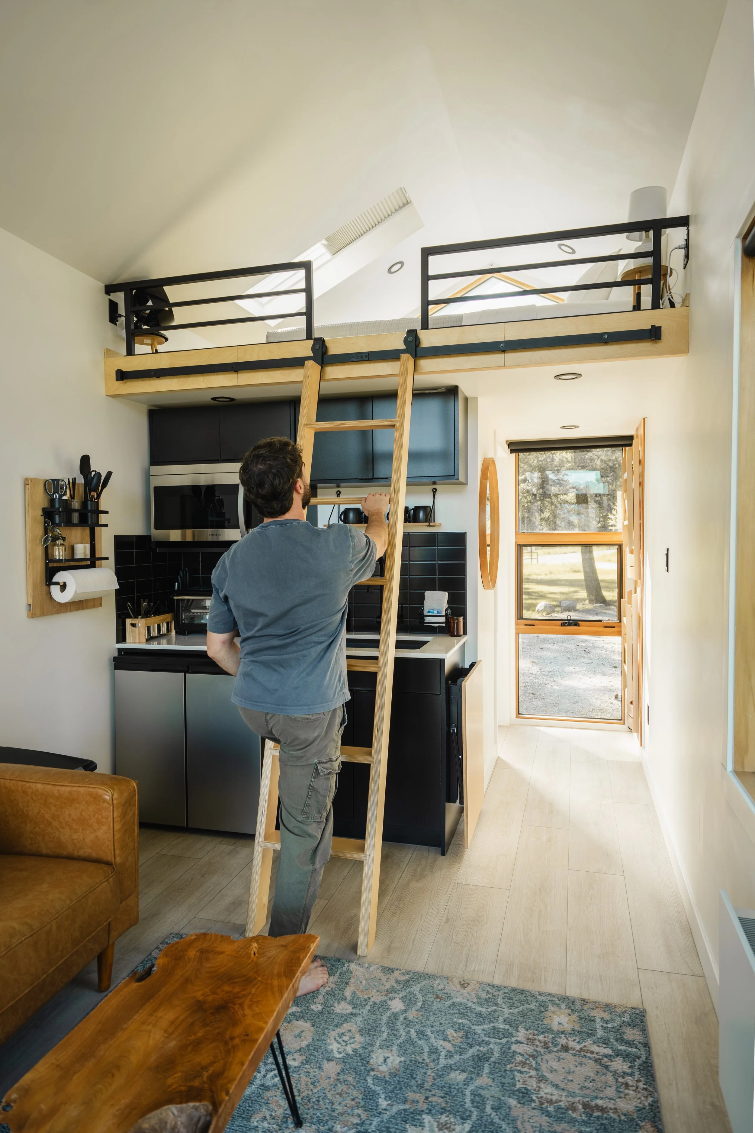 A man climbing a wooden ladder to access a loft bed in a modern kitchen and living space.
