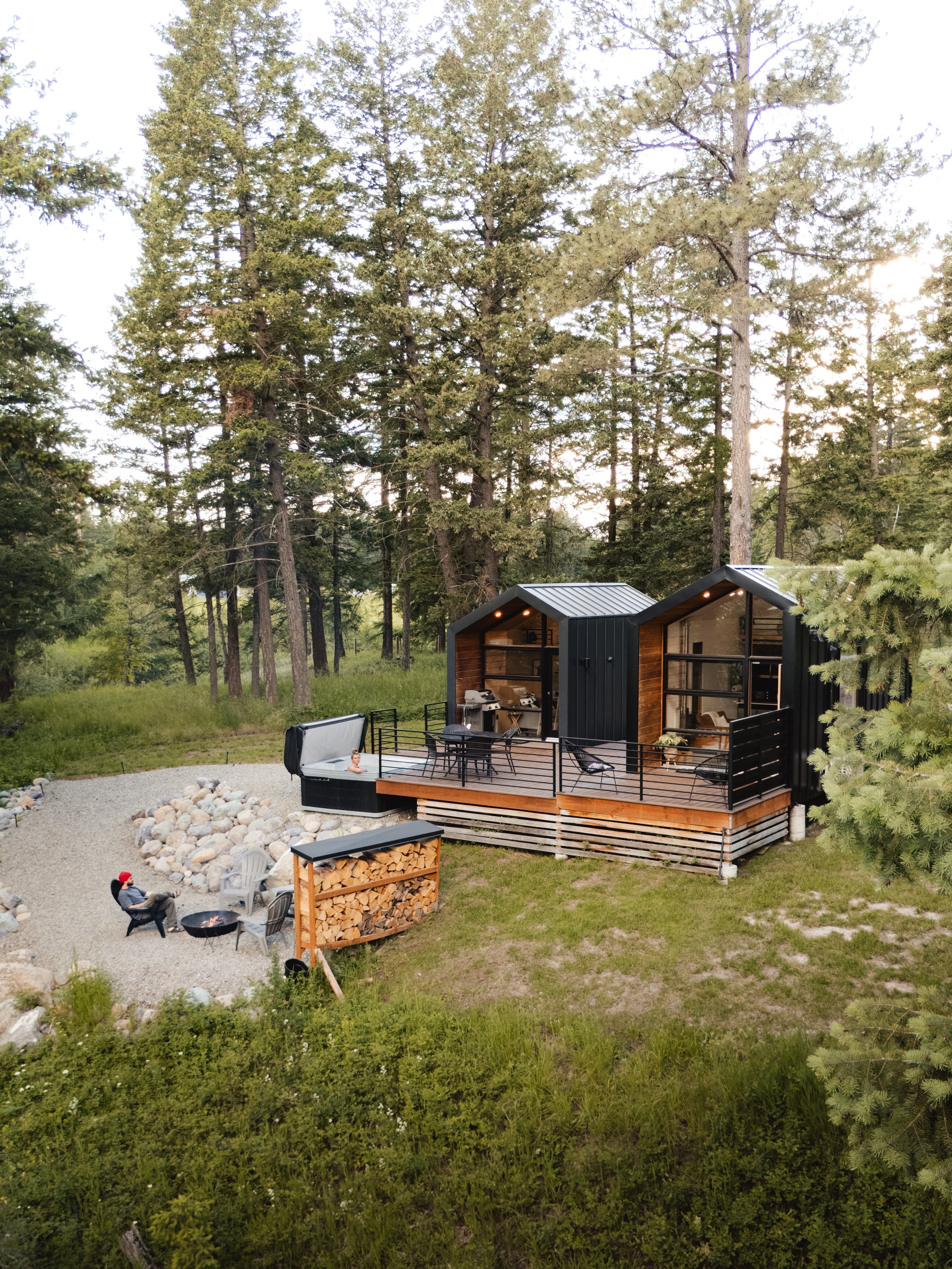 A cabin in the woods with a deck, outdoor seating, a fire pit, and tall trees surrounding the area.