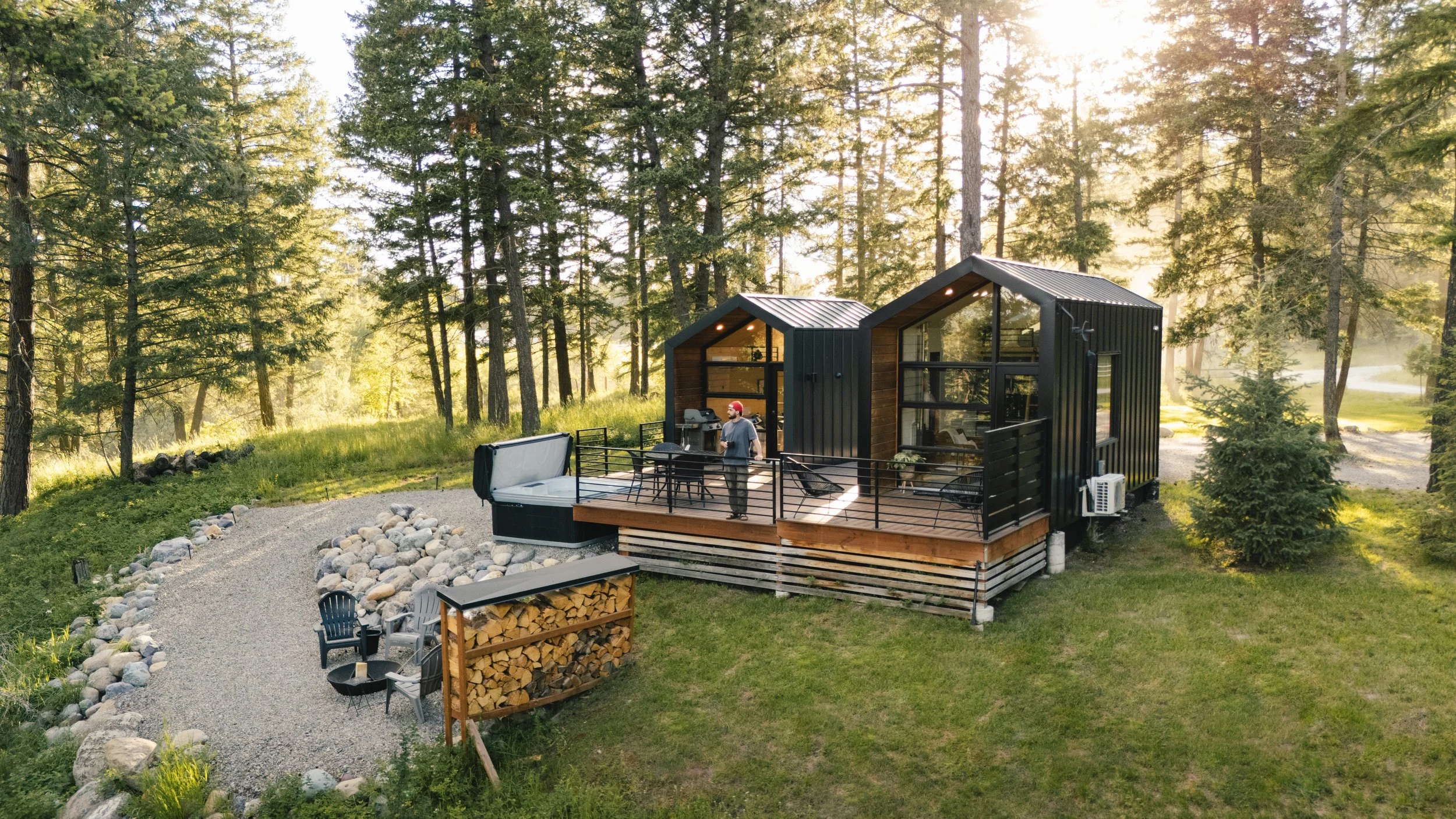 A modern tiny home with a black exterior and large windows, situated in a forested area during sunset. The house has a deck with outdoor furniture and a person standing on it, grilling. There is a firewood storage and a gravel pathway leading to the 