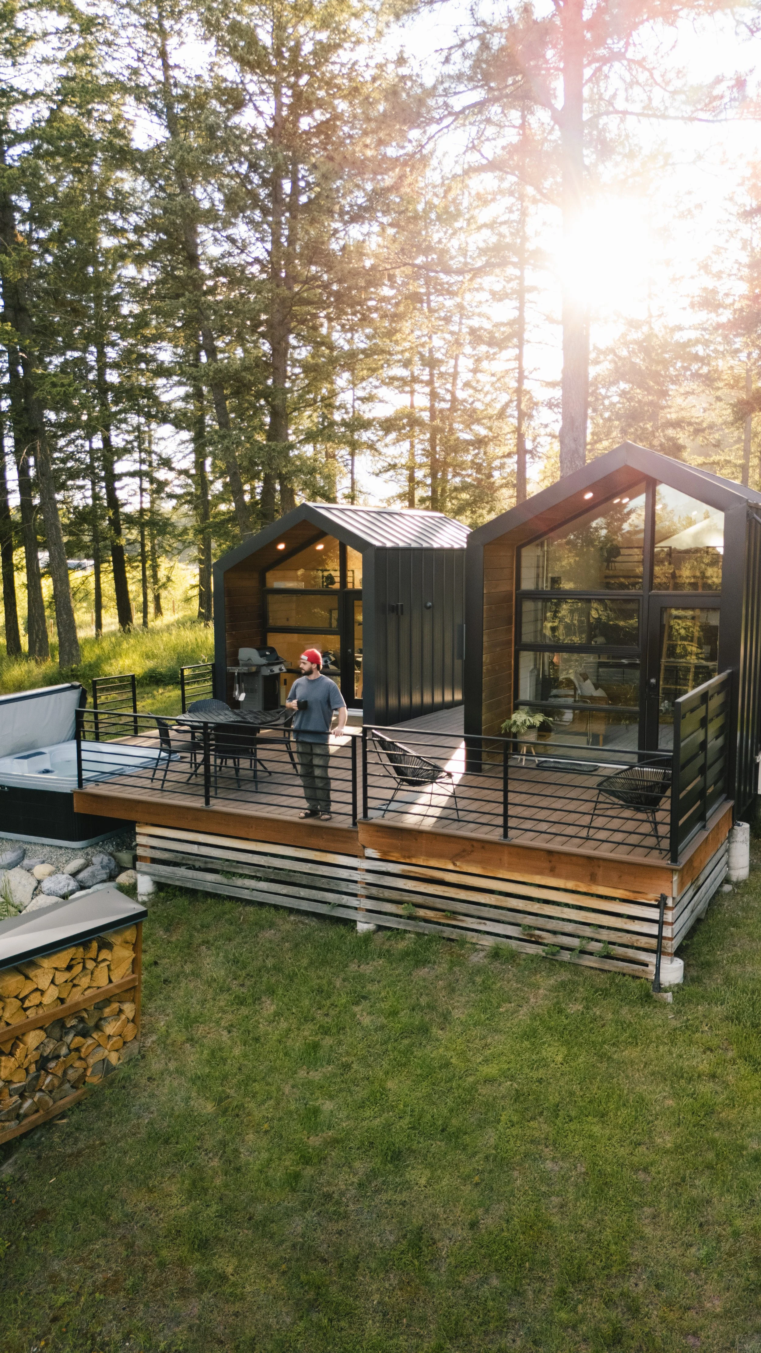 A man wearing a red cap and gray t-shirt standing on a wooden deck with black railing in a wooded area with tall pine trees, sun shining through the trees, and two small modern cabins with large glass windows and black metal roofs.
