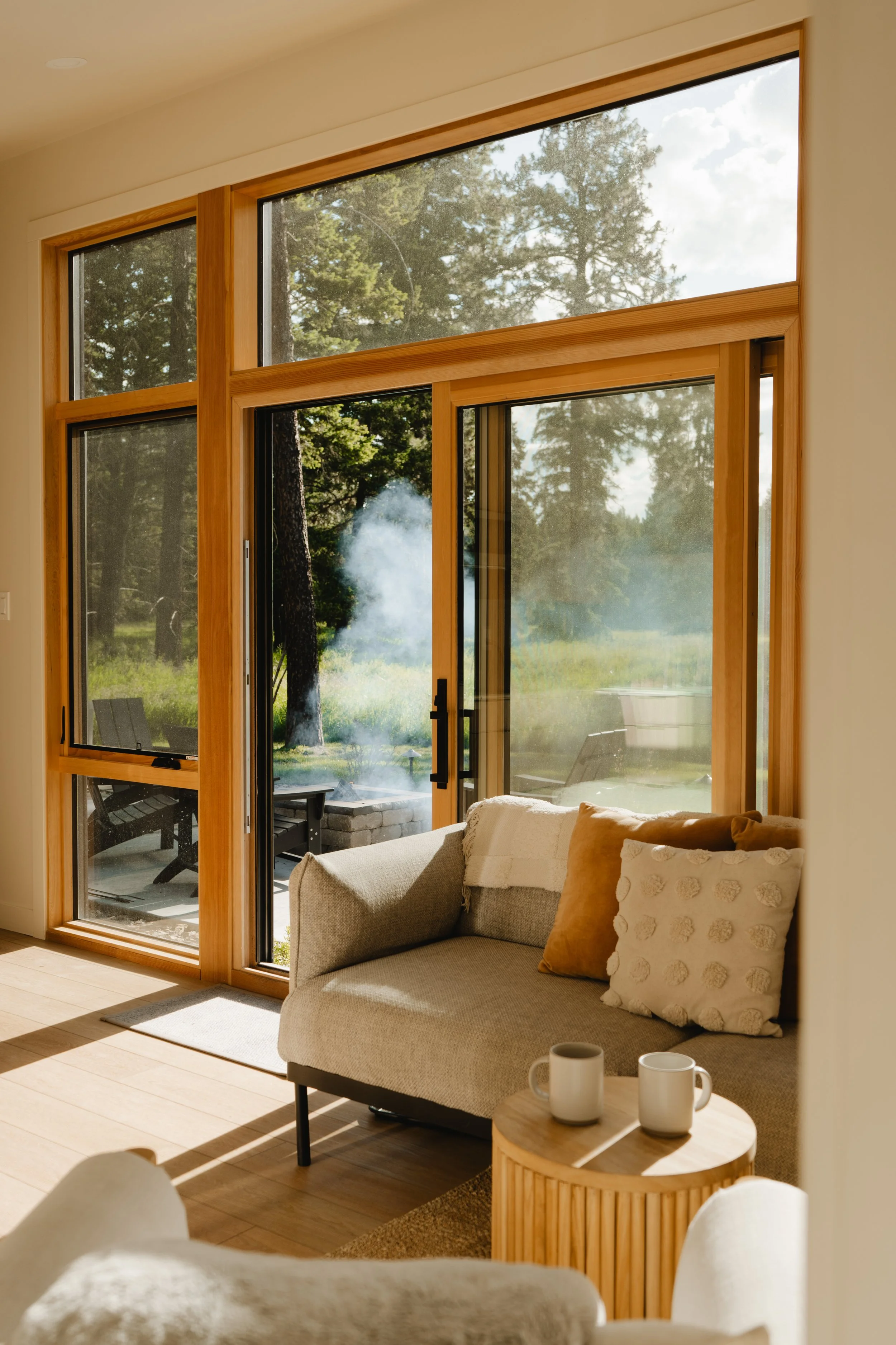 Living room with large wooden framed glass sliding doors leading to an outdoor patio, a beige couch with decorative pillows, a round wooden side table with two white mugs, and a view of trees and sky outside.