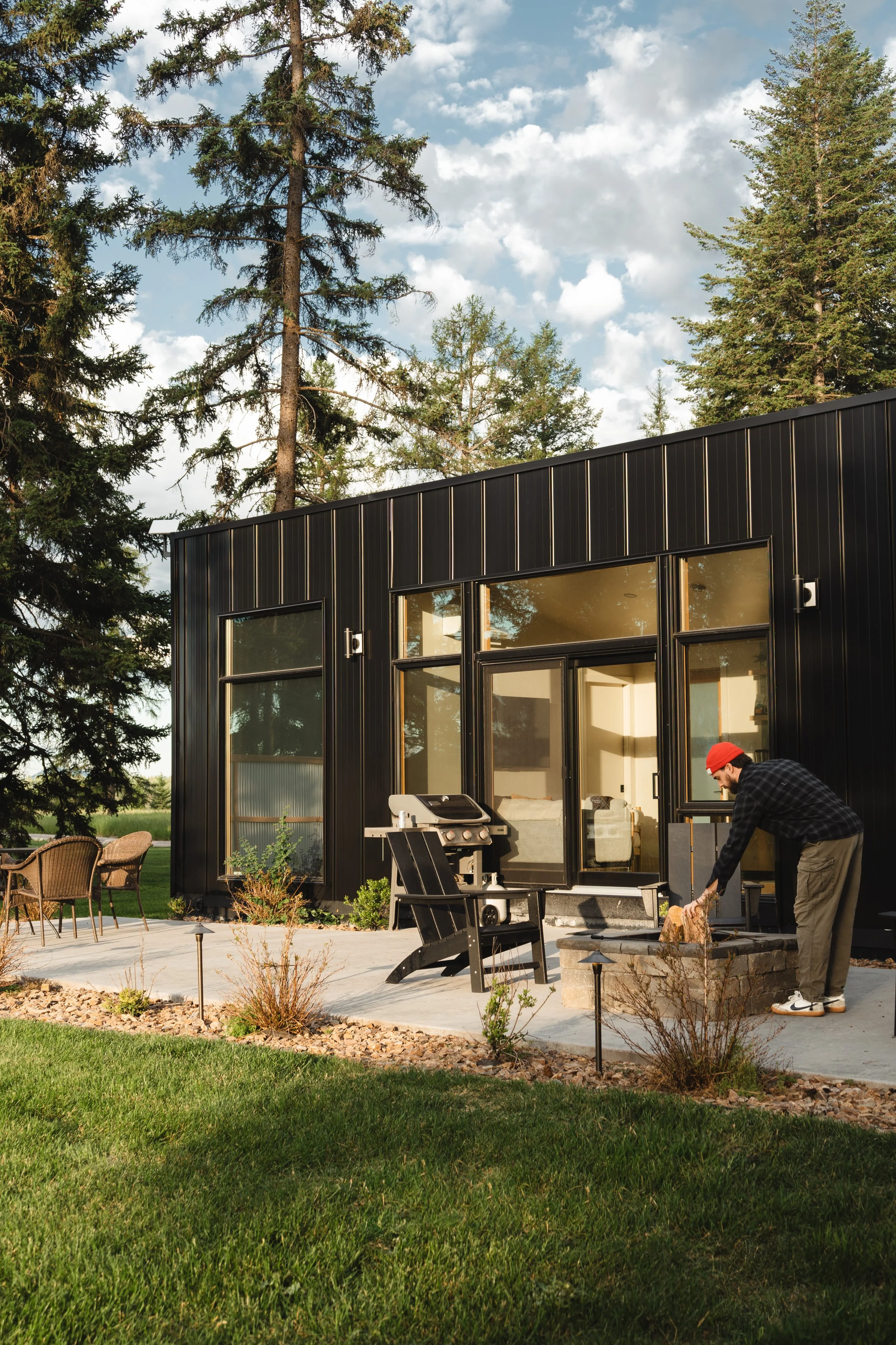 A man wearing a red cap and black jacket is kneeling in a backyard, stacking wood logs in front of a modern black metal house with large windows. There are patio furniture and a barbecue grill visible, with trees and a partly cloudy sky in the backgr