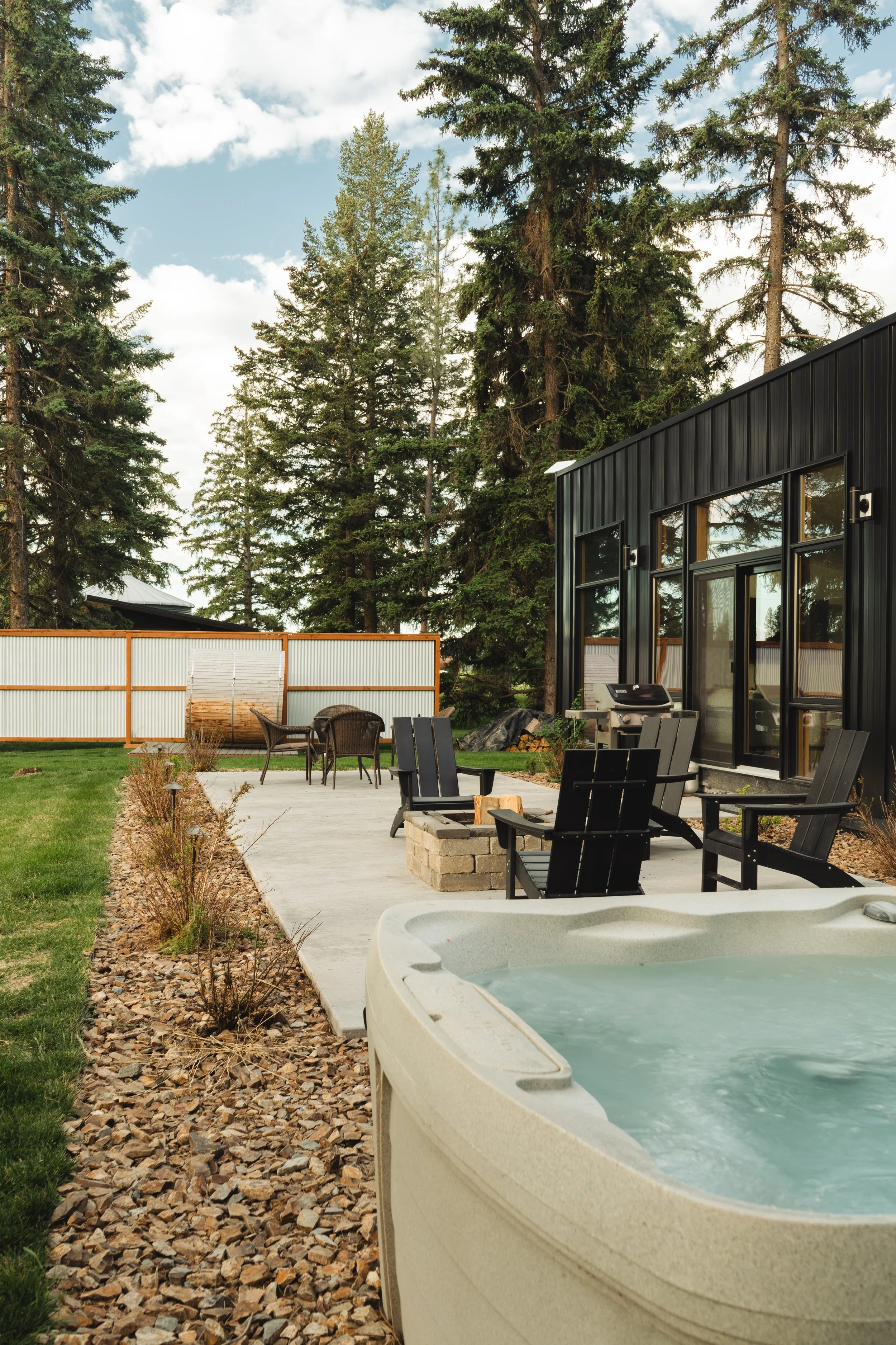 Outdoor backyard space with a hot tub, patio area with chairs and a fire pit, tall trees, and a modern black house with large windows.