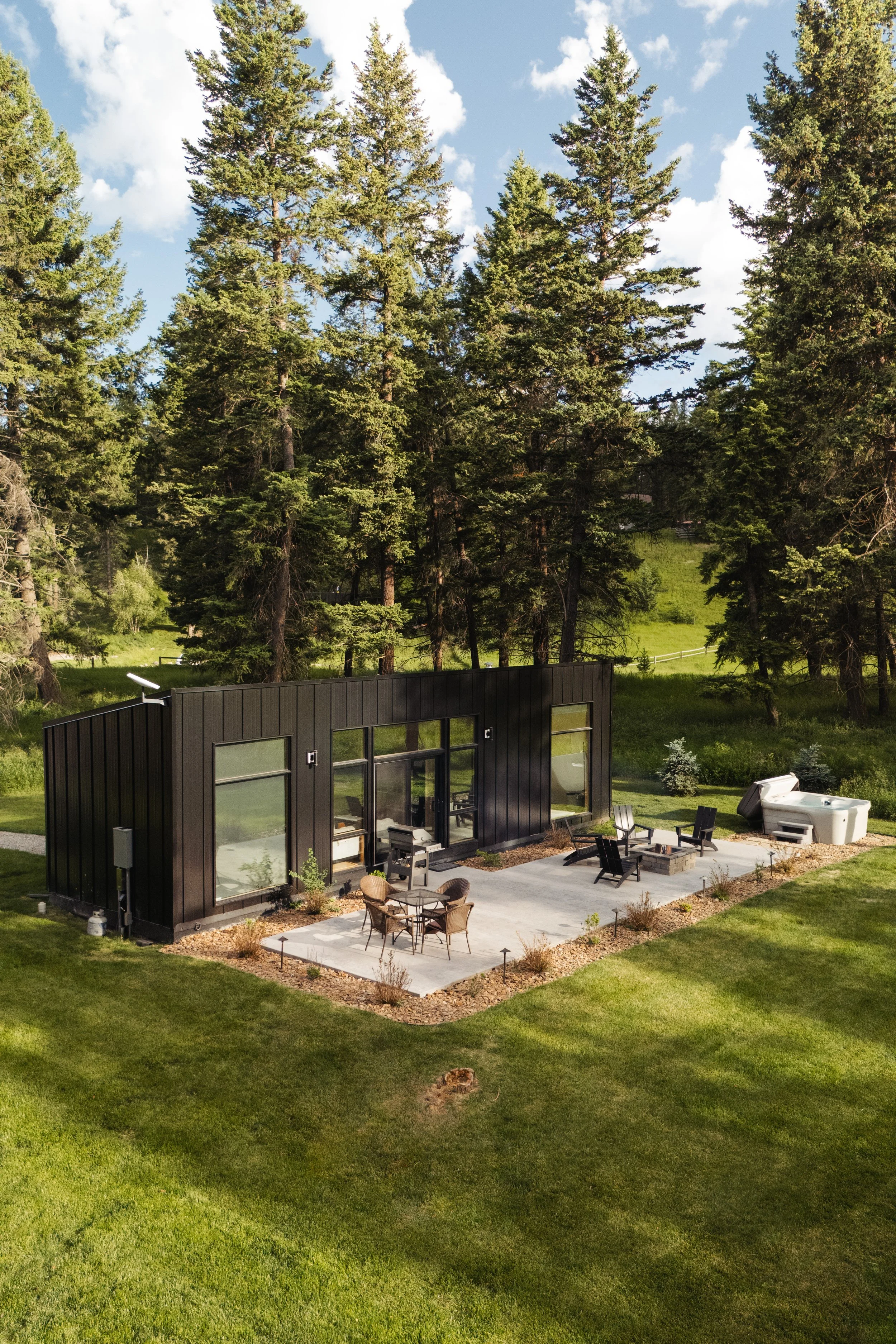 Modern black house with large glass windows, outdoor patio with seating, in a lush green yard surrounded by tall pine trees on a clear day.