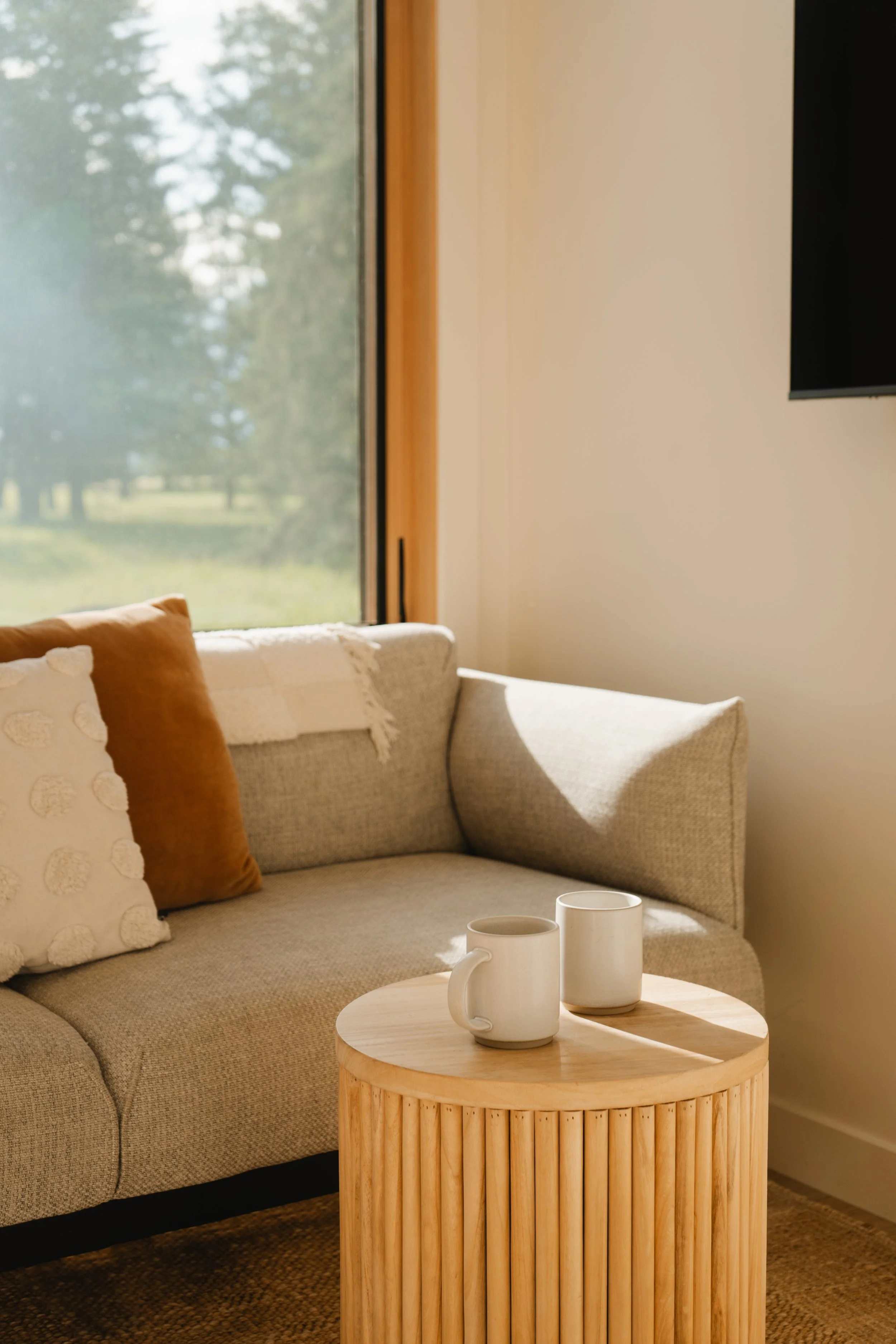 Living room with beige couch, orange and white pillows, a wooden side table with two white mugs, large window showing trees outside, and a wall-mounted TV.