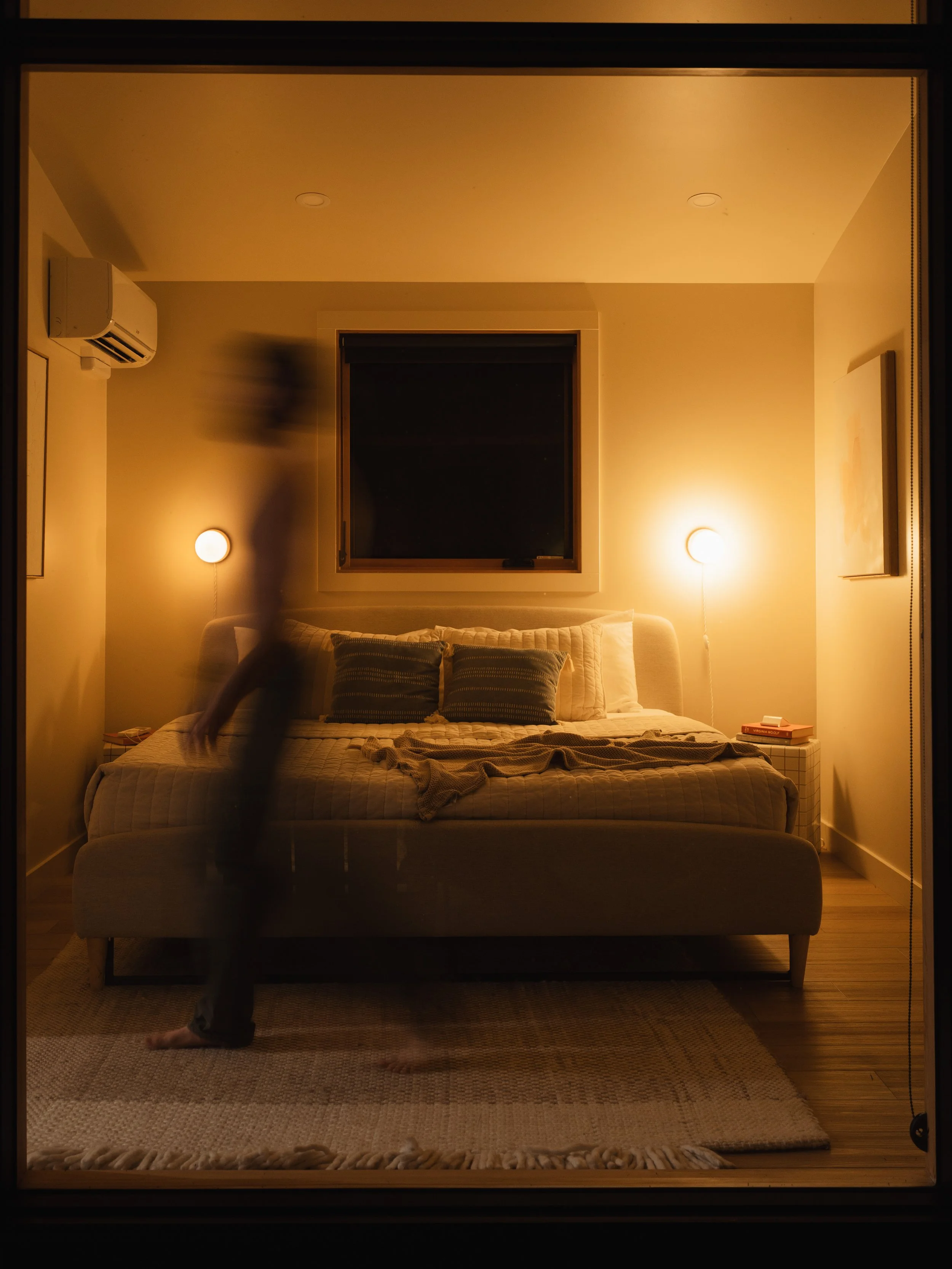A cozy bedroom with warm lighting, a large bed with multiple pillows, a window with closed blinds, and an outdoor scene with a house and trees visible at night. A person is walking past the bed, captured in motion.