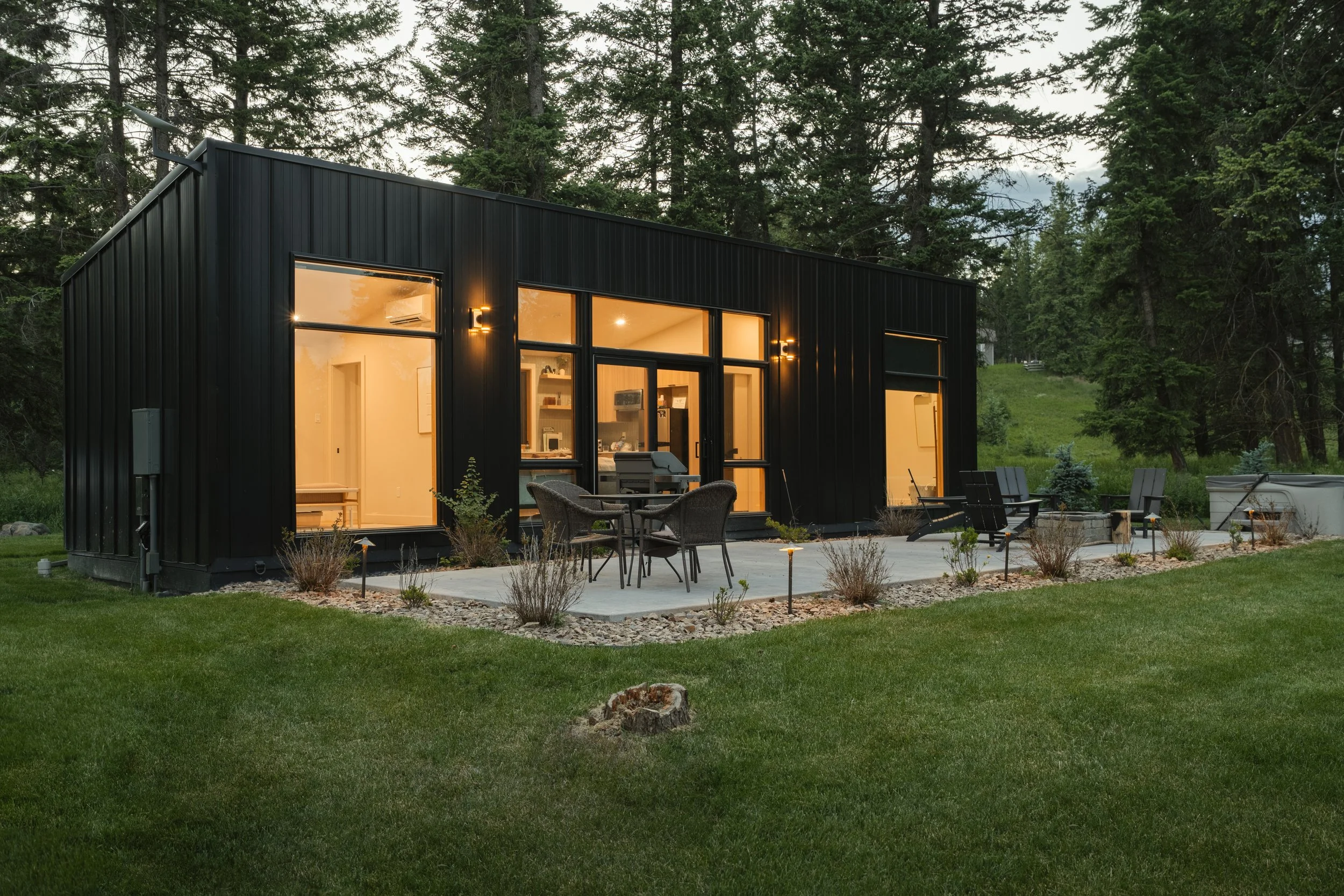 Modern black house with large windows illuminated from inside, surrounded by a grassy backyard with outdoor seating and a firepit, set in a wooded area during dusk.