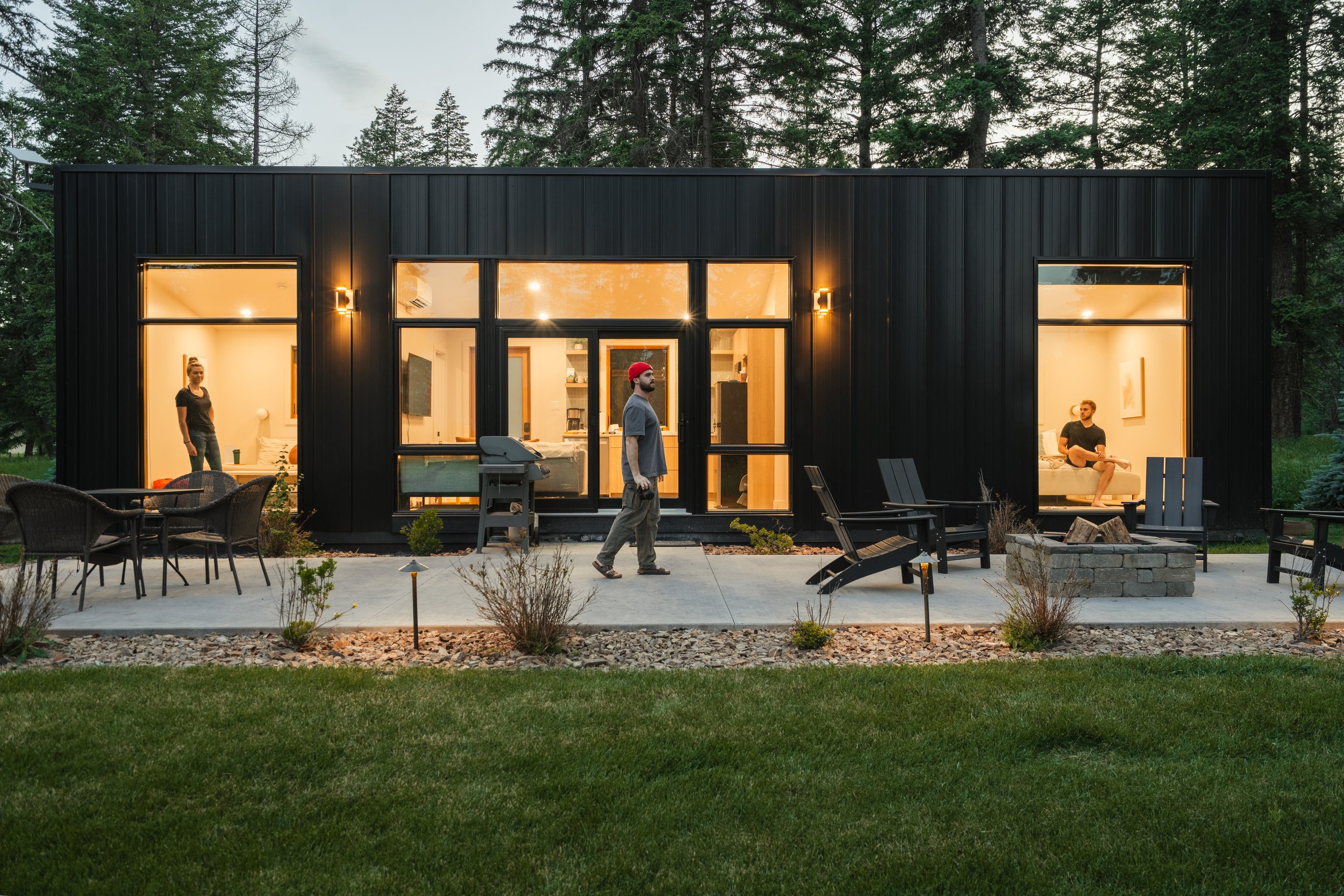 Modern black house with large glass windows and doors, illuminated from inside, with three people (a woman and two men) outside on a patio surrounded by trees.