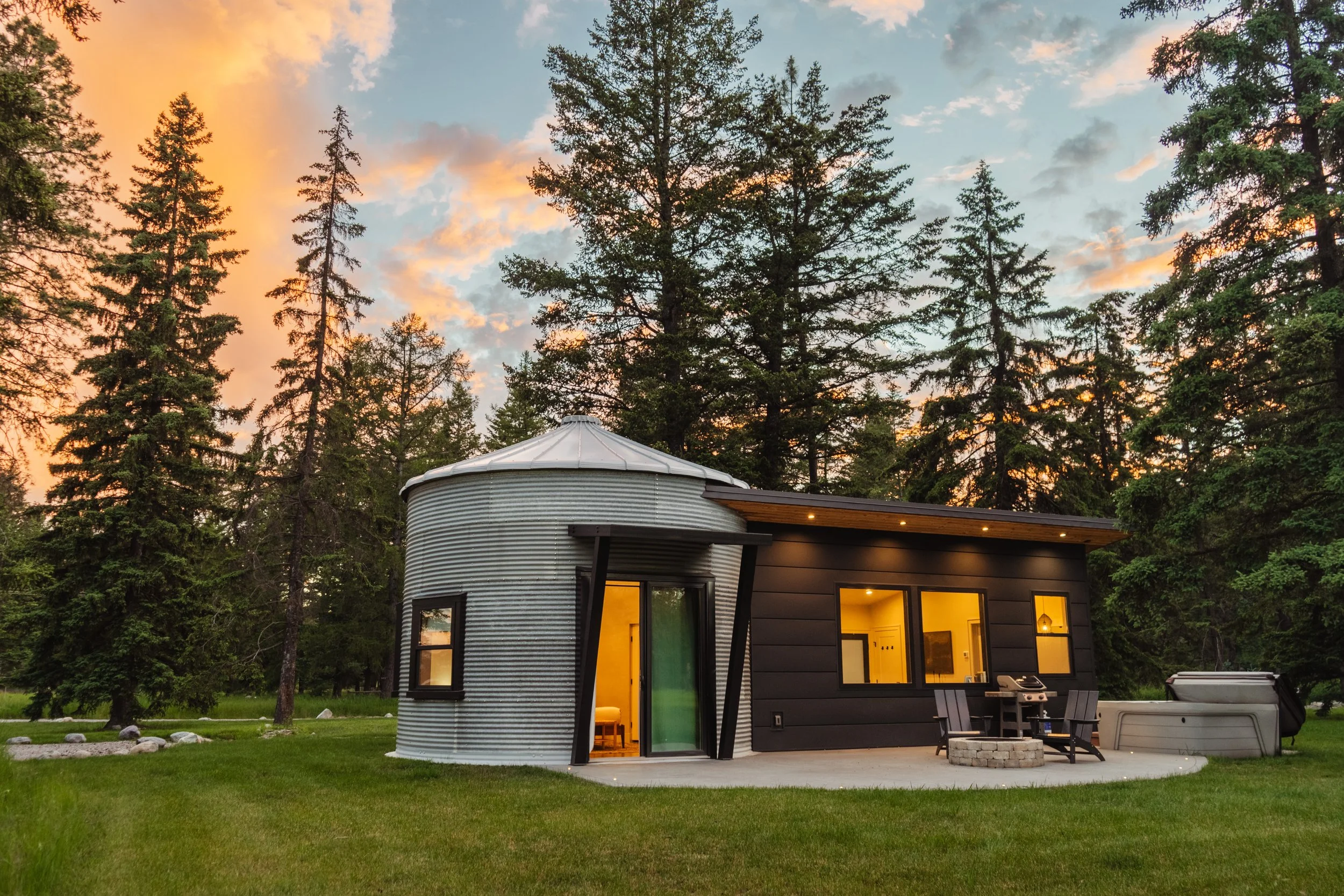 A modern tiny house with a black exterior and metal siding, surrounded by green lawn and tall pine trees, under a sky with clouds during sunset. Lights inside are on, and an outdoor patio with chairs and a grill is visible.