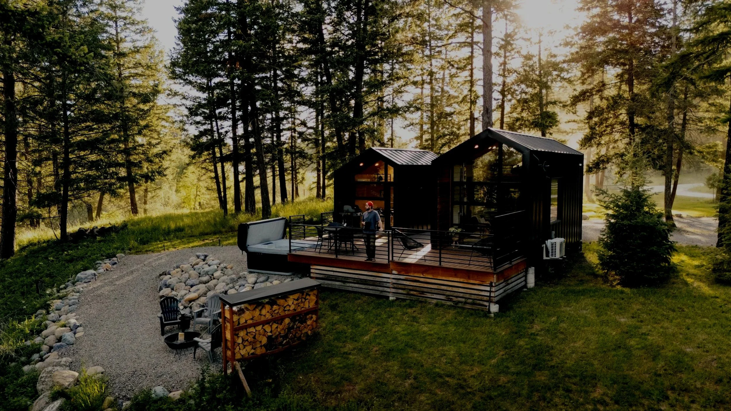 A modern black cabin with large glass windows in a forested area during sunset, featuring a wooden deck with outdoor furniture and a man standing on it.