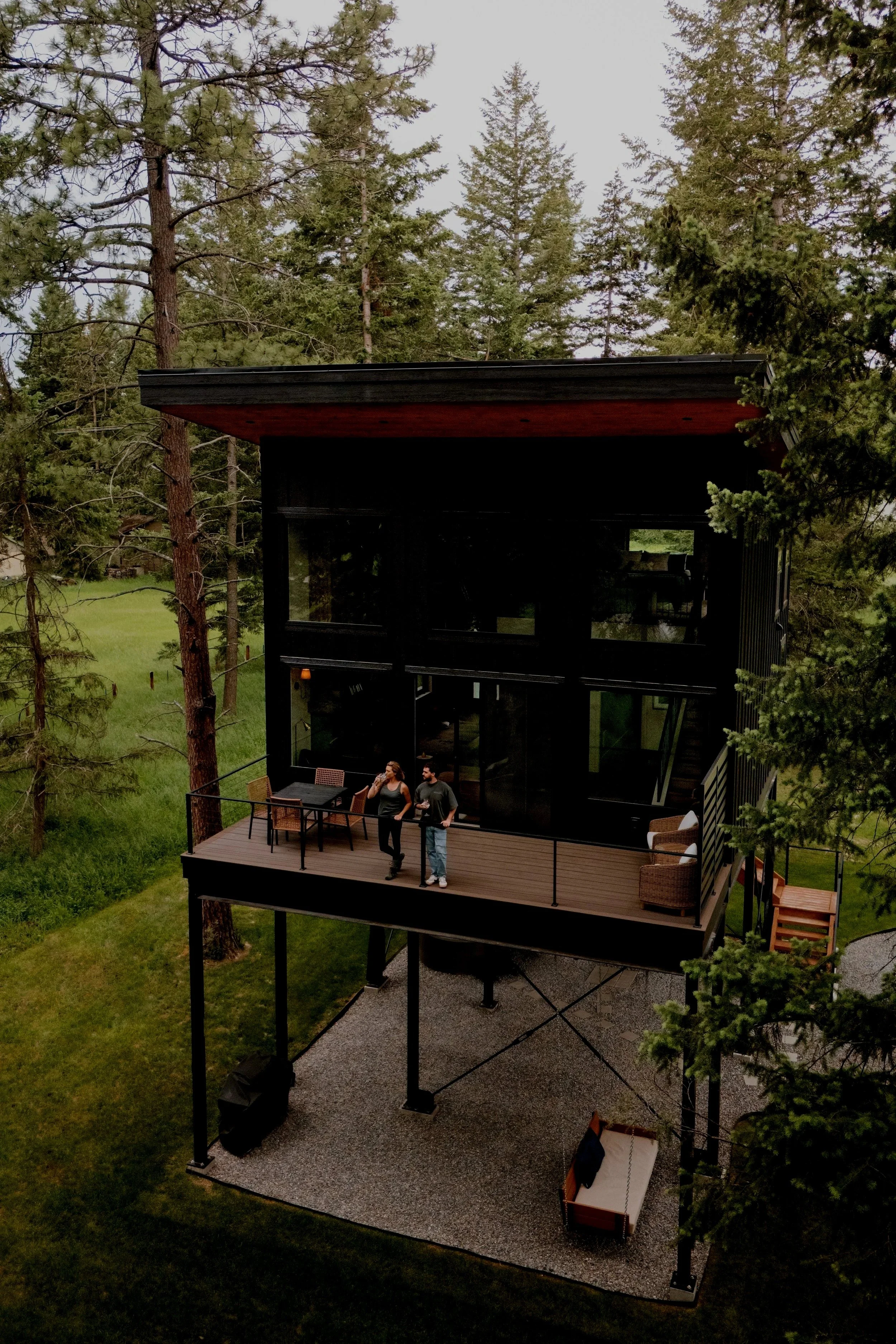 A modern house on stilts situated among tall pine trees with a large deck. Two people are standing and talking on the deck, one holding a drink. The area below the deck is covered with gravel, and there is an outdoor swing on the right.