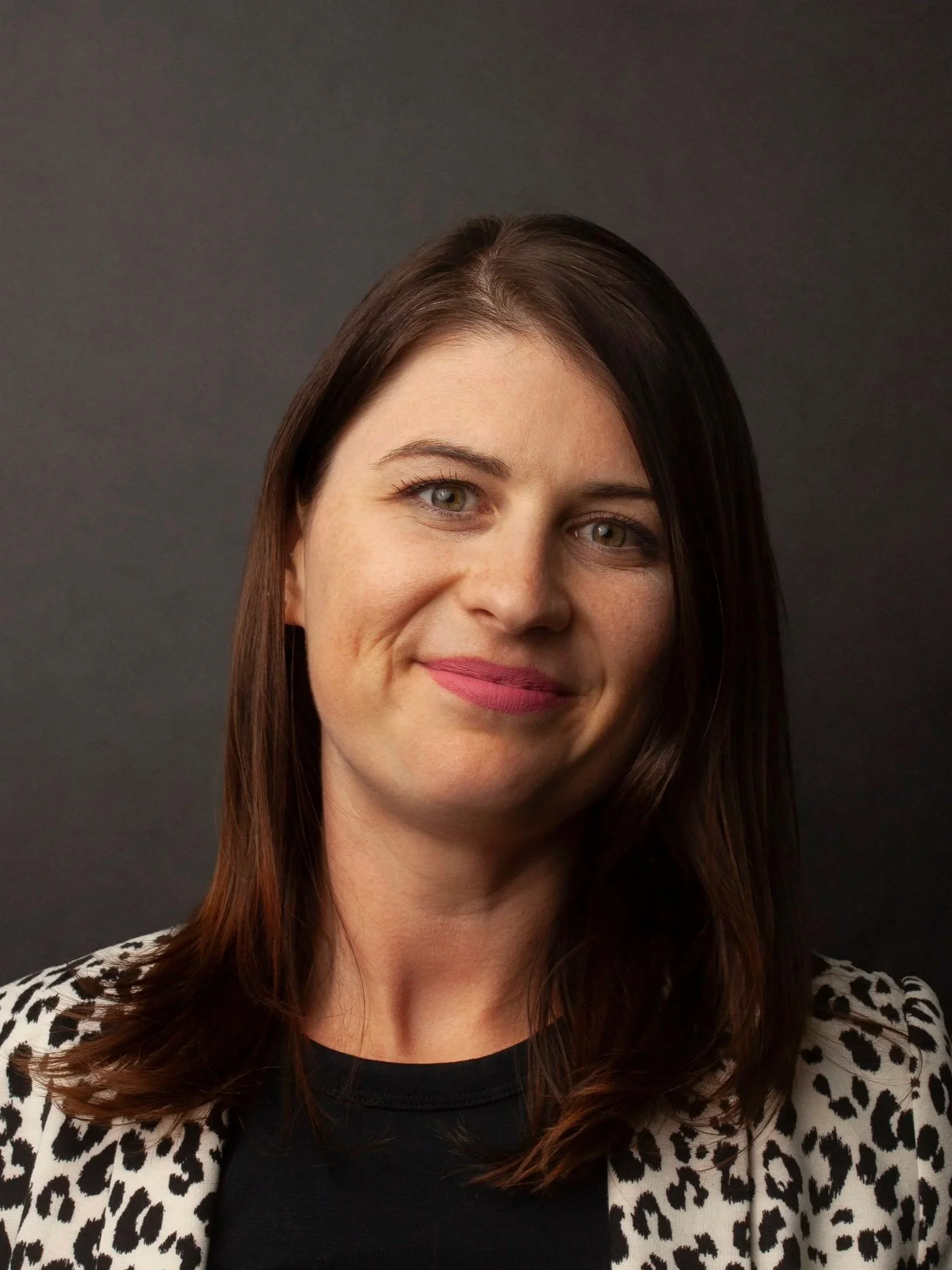 Portrait of a female CPG Shopper Marketing Expert with brown hair, blue eyes, wearing a black top and a white leopard print jacket, smiling against a dark background.