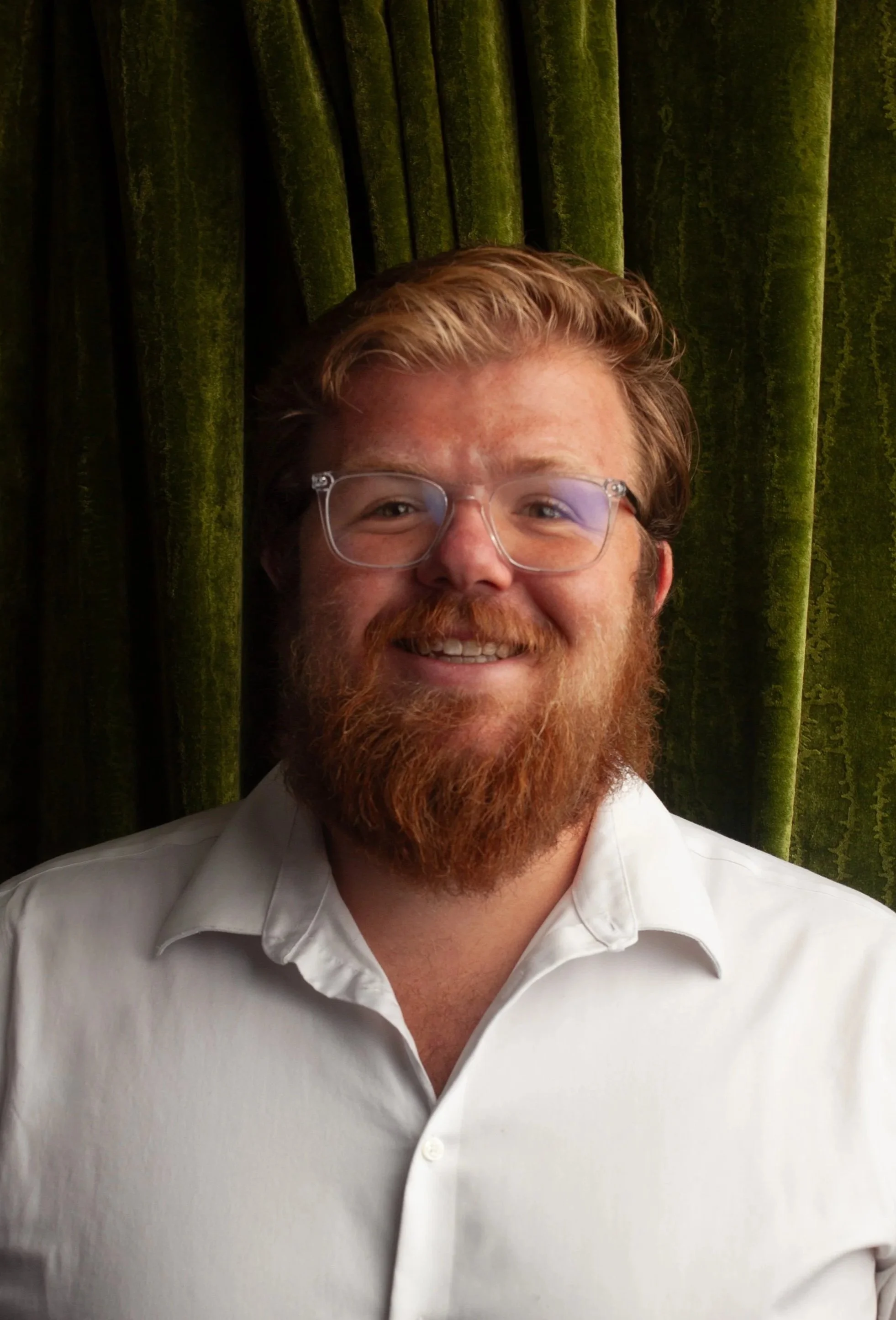 A smiling male CPG Shopper Marketing Expert with glasses and a beard, wearing a white shirt, standing in front of a green curtain.