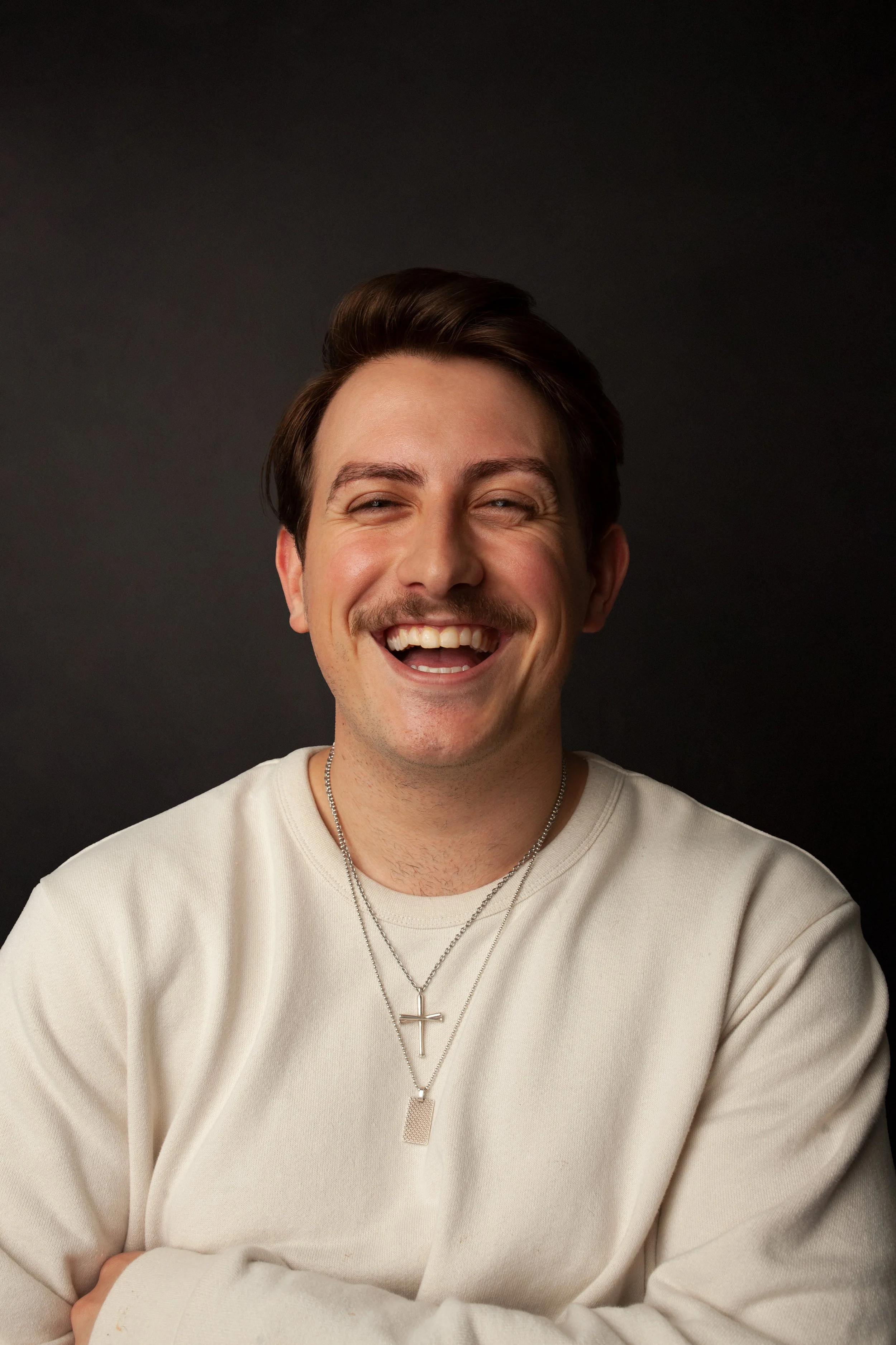 A young male CPG Shopper Marketing Expert with brown hair and a mustache, smiling and wearing a beige sweater with layered necklaces featuring cross and rectangular pendants, against a black background.