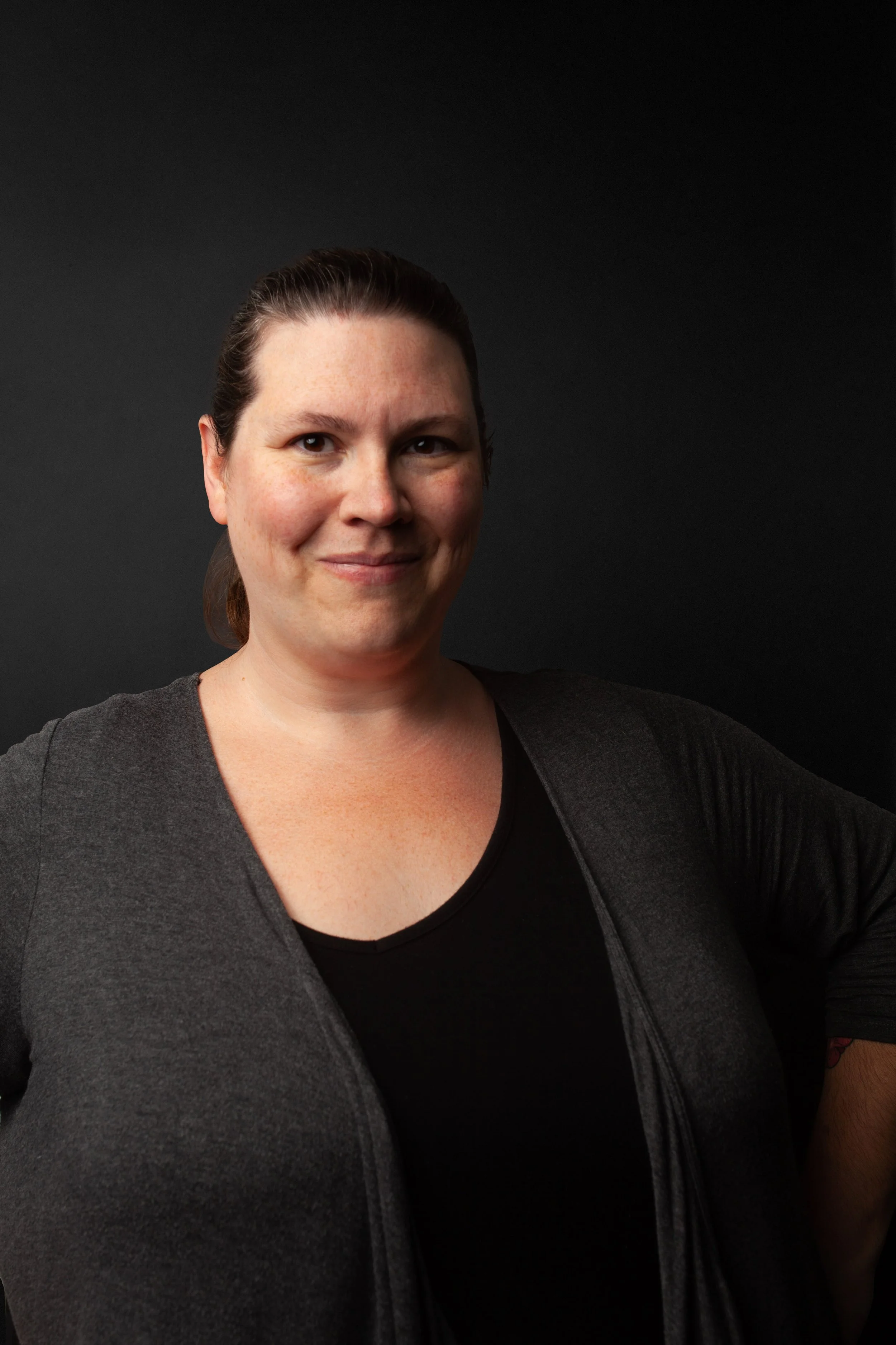 Portrait of a female CPG Shopper Marketing Expert with dark hair pulled back, wearing a black top and a dark gray cardigan against a dark background, smiling slightly.