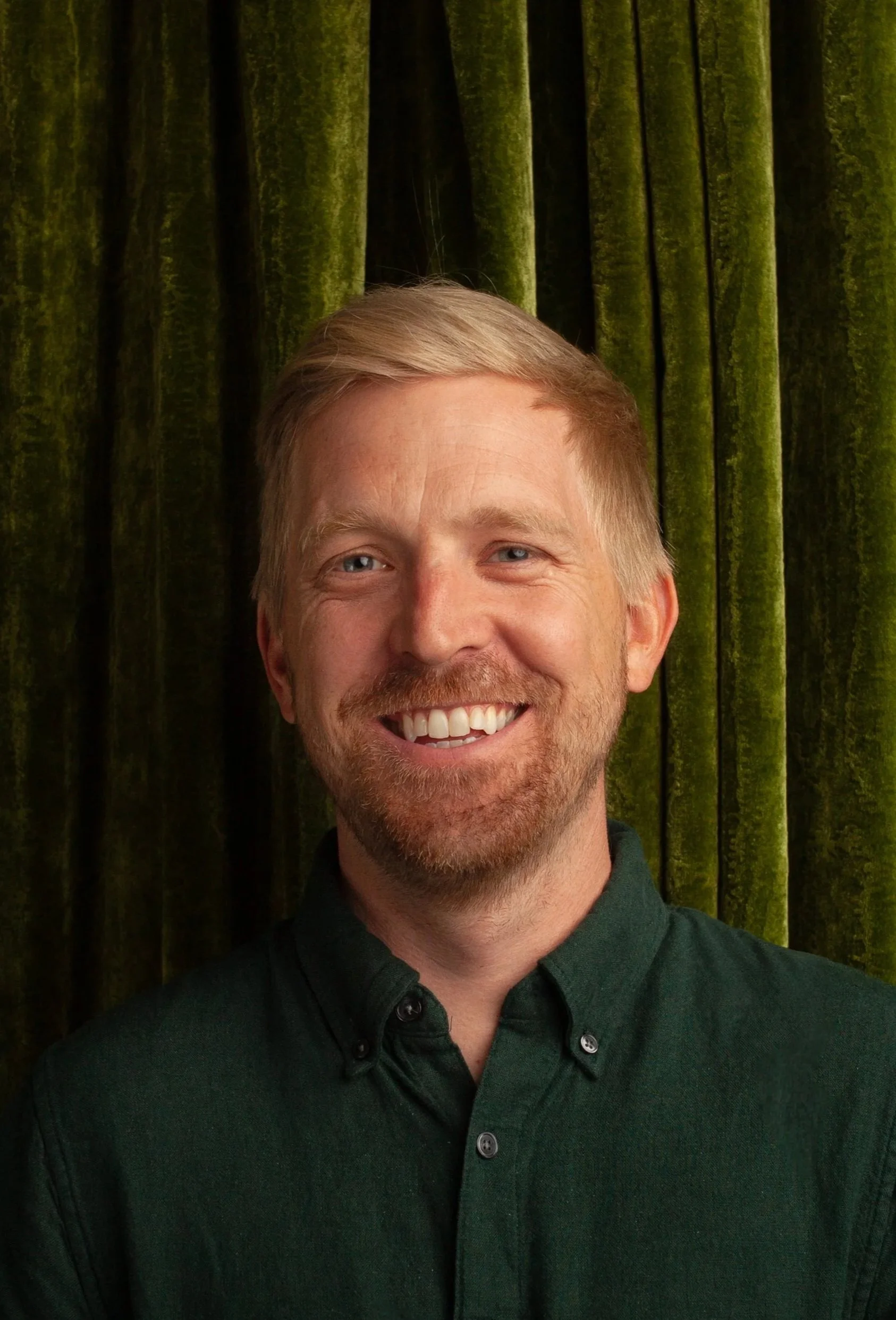 A smiling CPG Shopper Marketing Expert with blond hair and a beard, wearing a dark green button-up shirt, in front of a background of green velvet curtains.