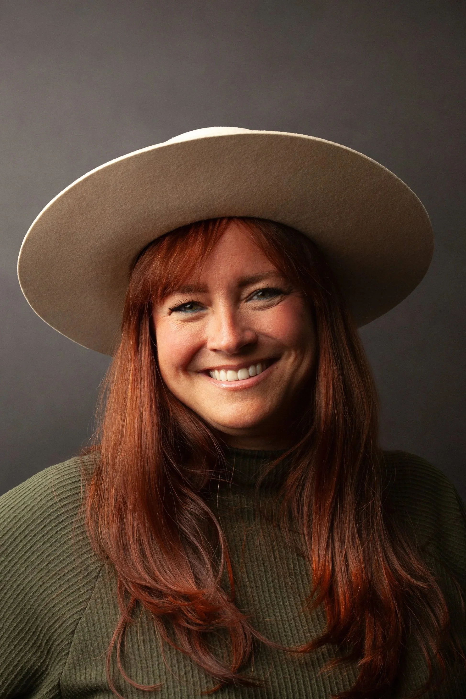 Close-up of a smiling female CPG Shopper Marketing Expert with long red hair wearing a wide-brimmed beige hat and a green ribbed top against a dark background.