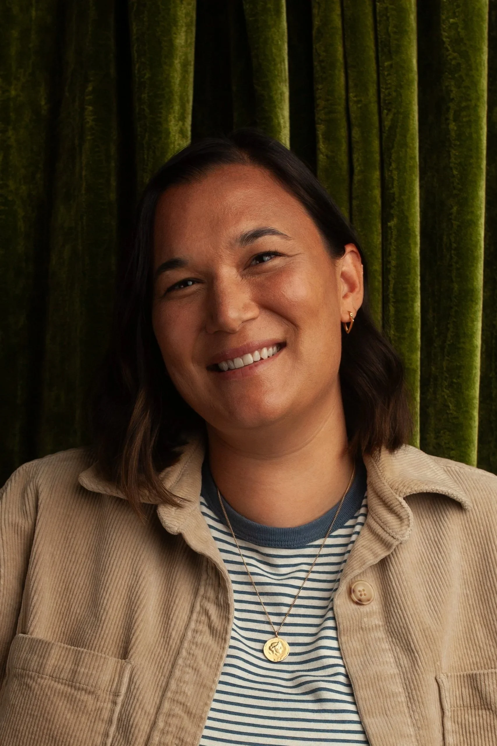 Smiling female CPG Shopper Marketing Expert with dark hair, wearing a beige jacket and striped shirt, standing in front of green velvet curtains.