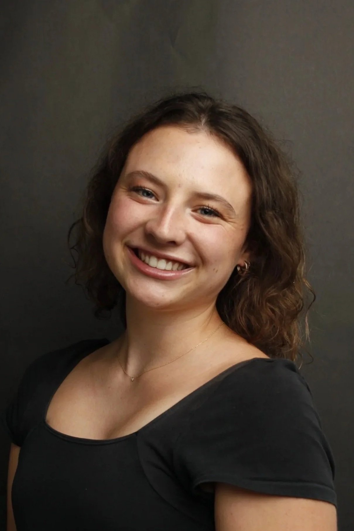 A young female CPG Shopper Marketing Expert with curly brown hair, fair skin, and blue eyes is smiling and wearing a black top with puffed sleeves, small earrings, and a delicate necklace. She stands against a dark background.