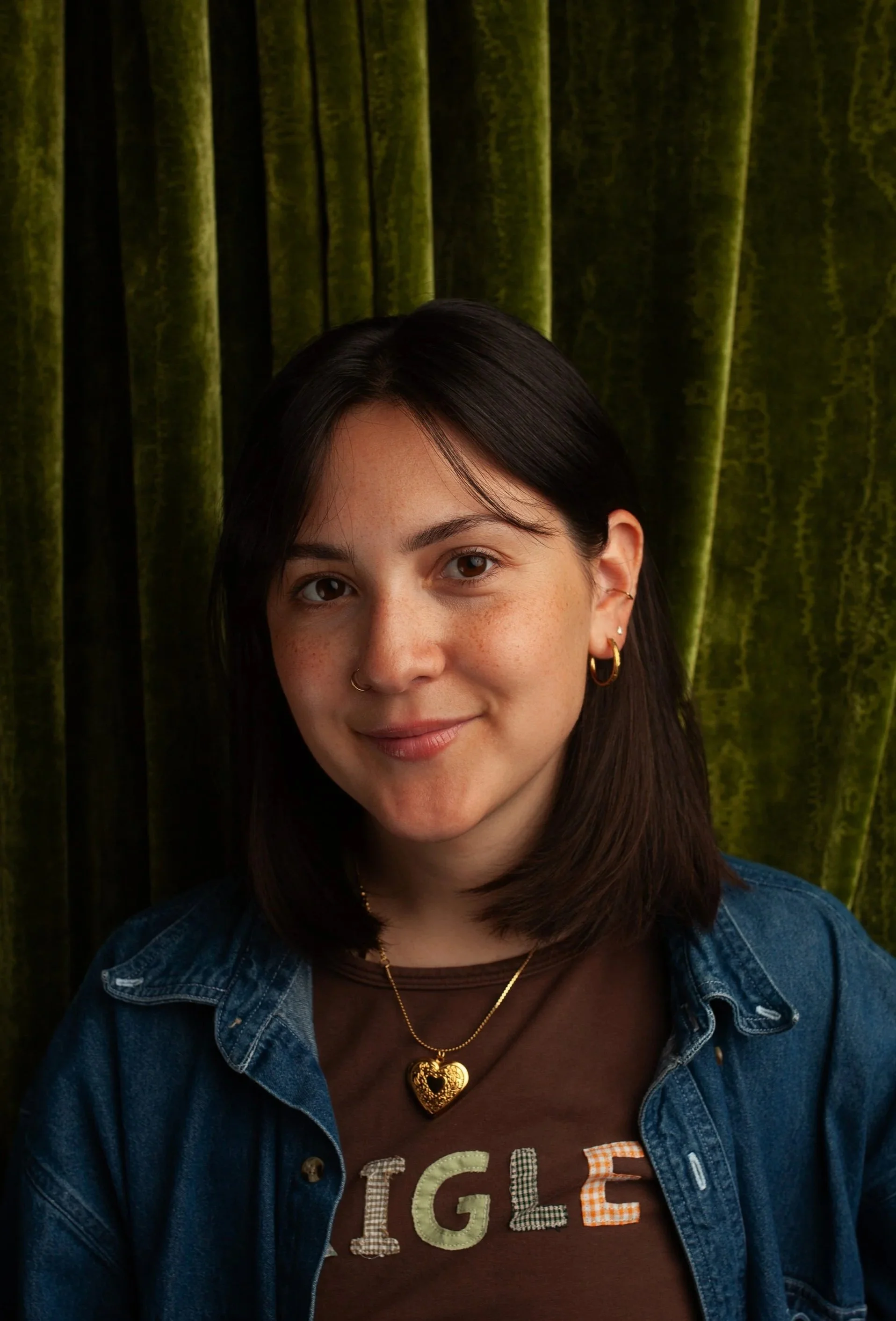 A young female CPG Shopper Marketing Expert with dark hair, wearing a denim jacket, a brown T-shirt with colorful lettering, a gold heart necklace, and multiple earrings, smiling in front of green velvet curtains.