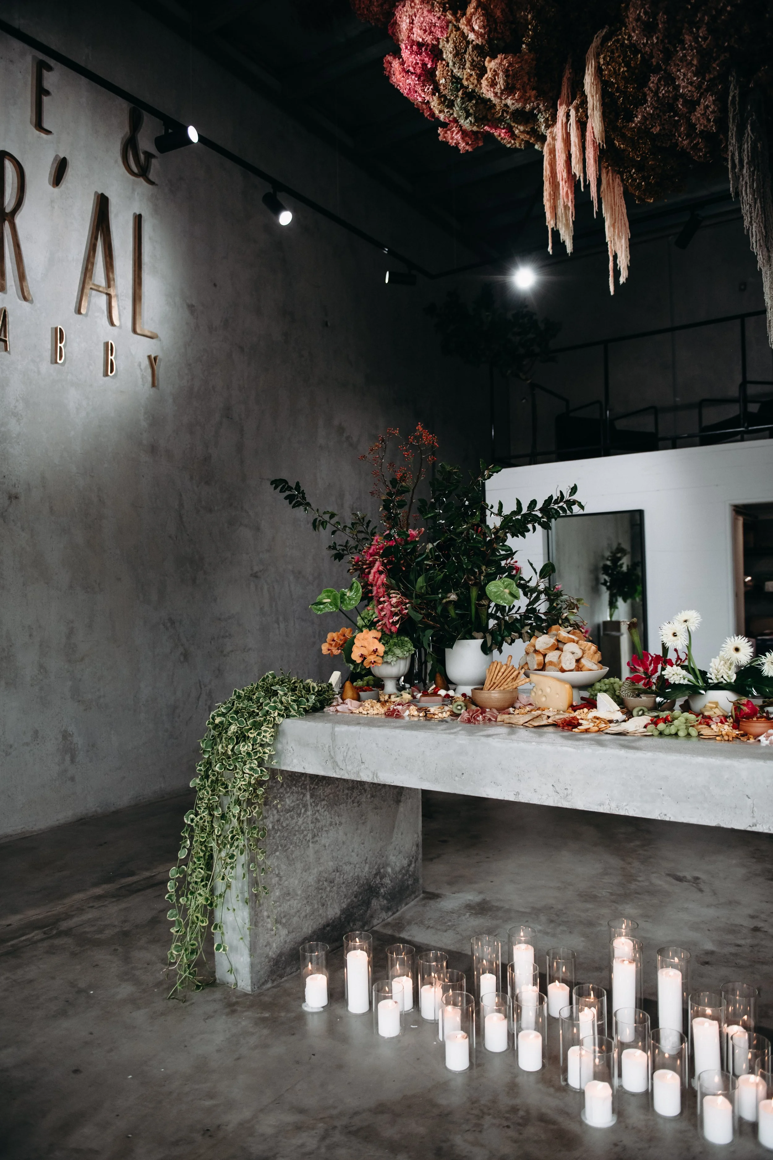 A modern, minimalist interior with a concrete table decorated with flowers, cheese, and snacks, and surrounded by white candles on the floor. The wall features large gold letters spelling 'esprial' and 'bakery.'
