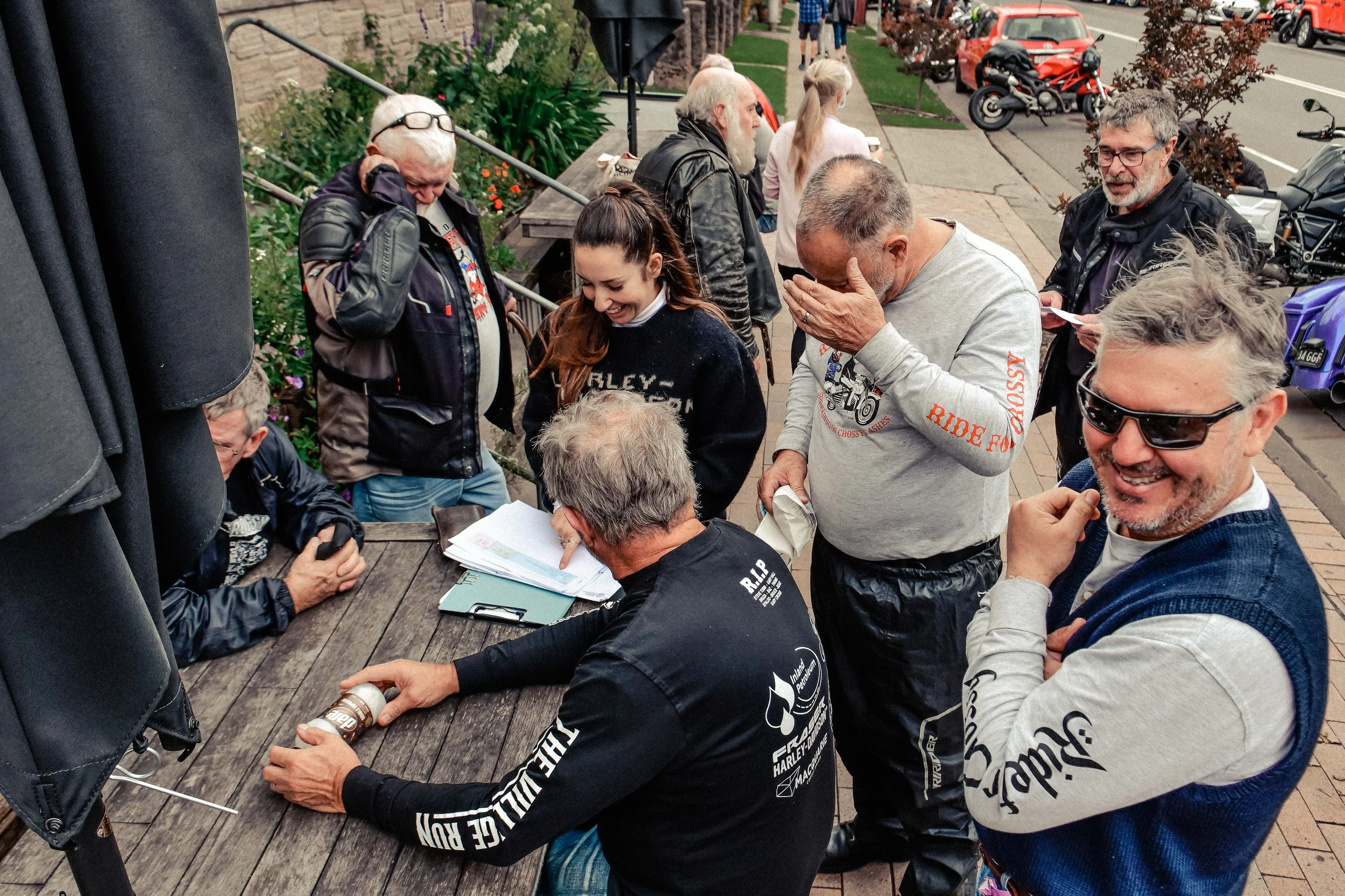 Group of people gathered around a wooden table outdoors, some looking at documents and chatting, with motorcycles and a street visible in the background.