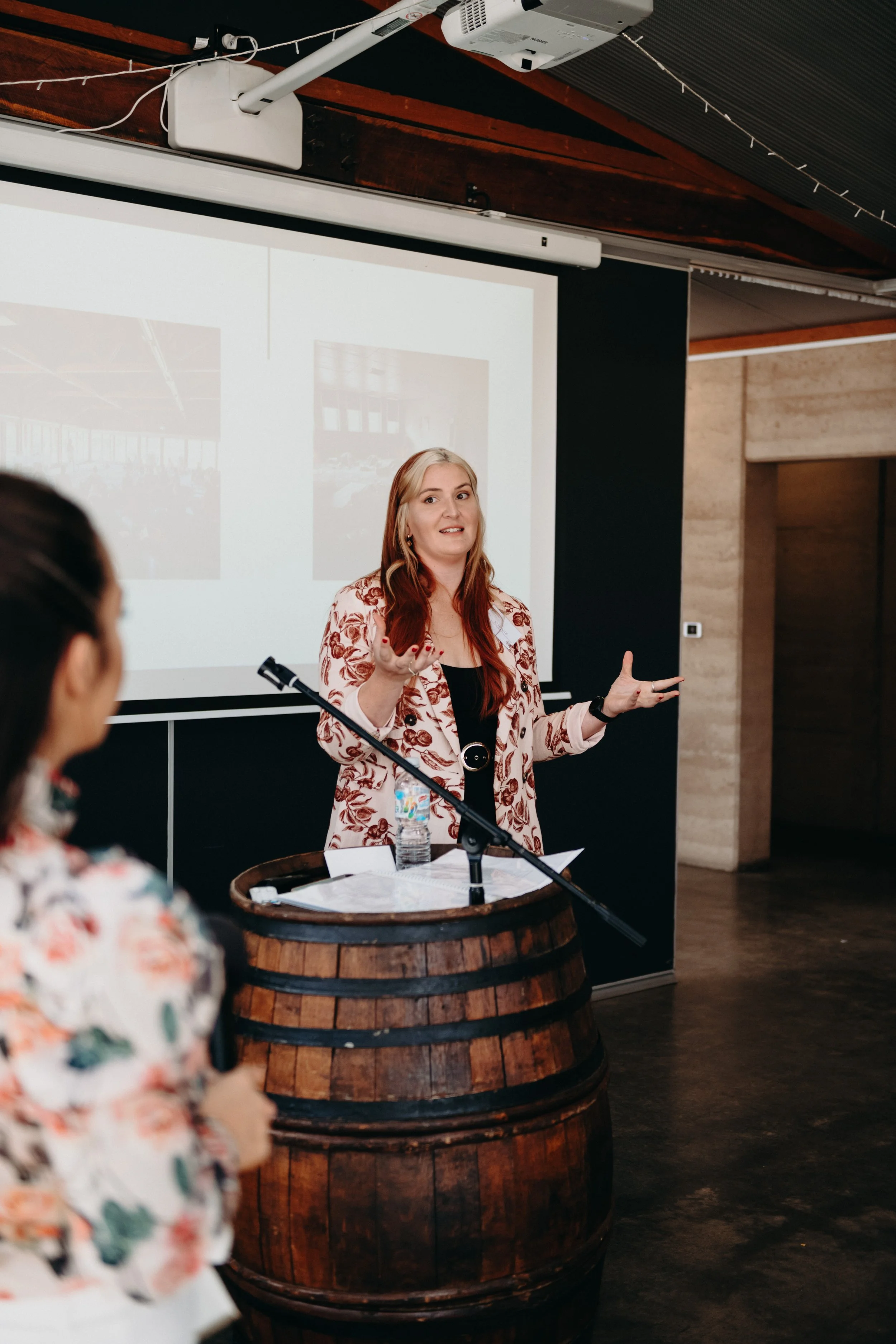 A woman giving a presentation in front of a projection screen with visual slides. She stands behind a wooden barrel, with notes and a water bottle on top, and gestures with her hands while talking.