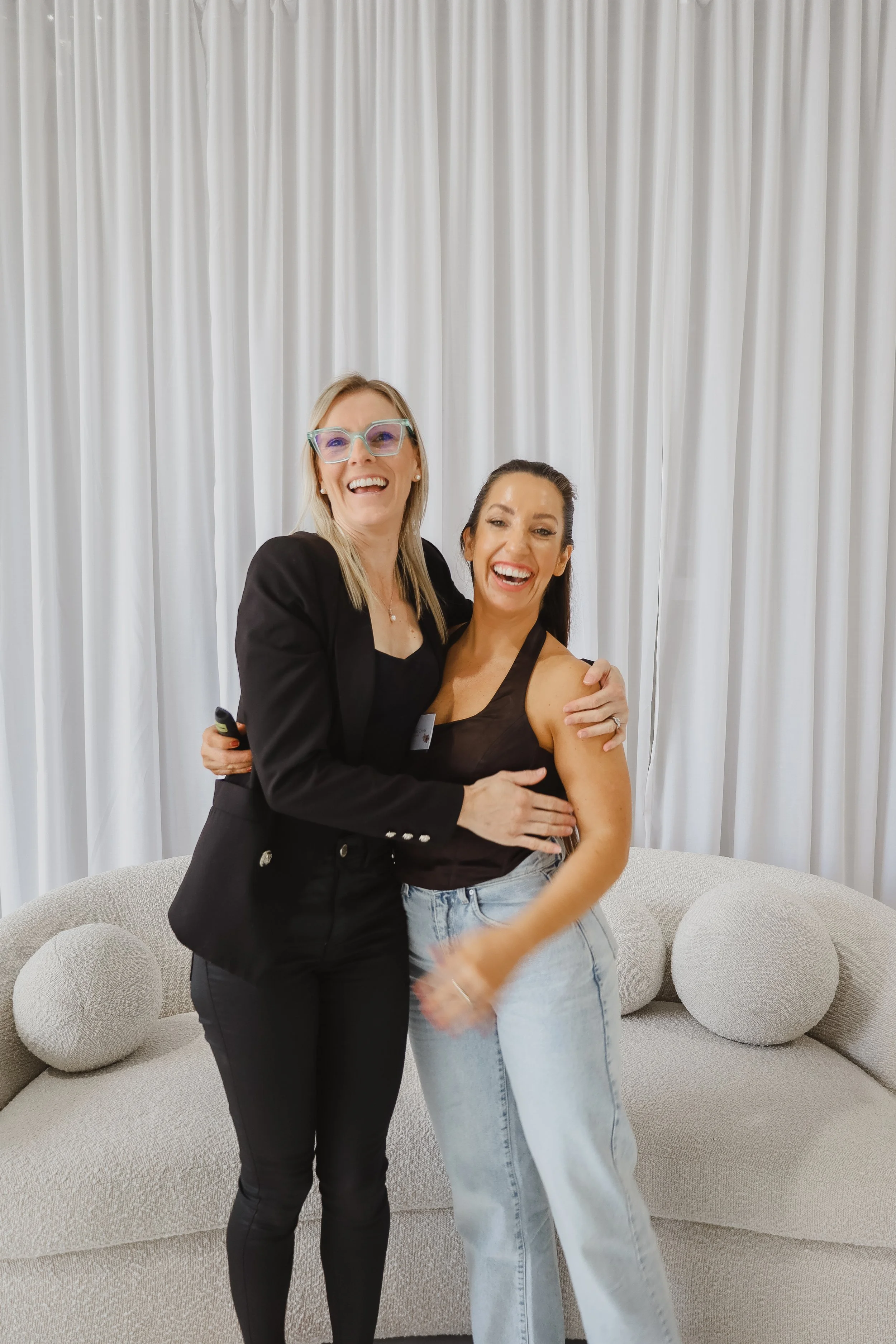 Two women hugging and smiling in front of a white curtain with a rounded beige sofa and decorative pillows behind them.