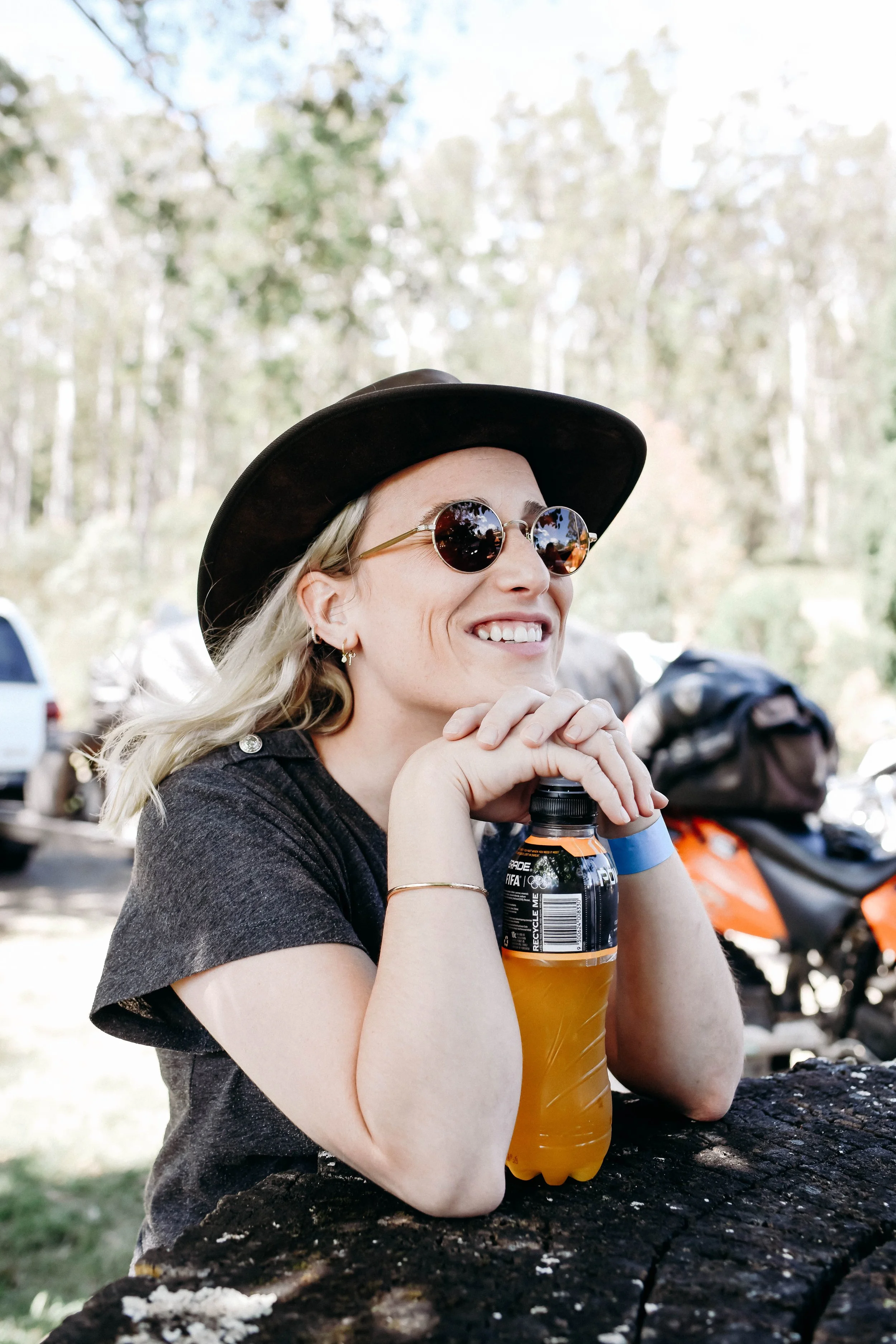 A woman with blonde hair, wearing a black cowboy hat and sunglasses, smiles while resting her chin on her hands, holding a bottle of orange sports drink. She is seated outdoors at a wooden table in a wooded area with parked cars and motorcycles in the background.