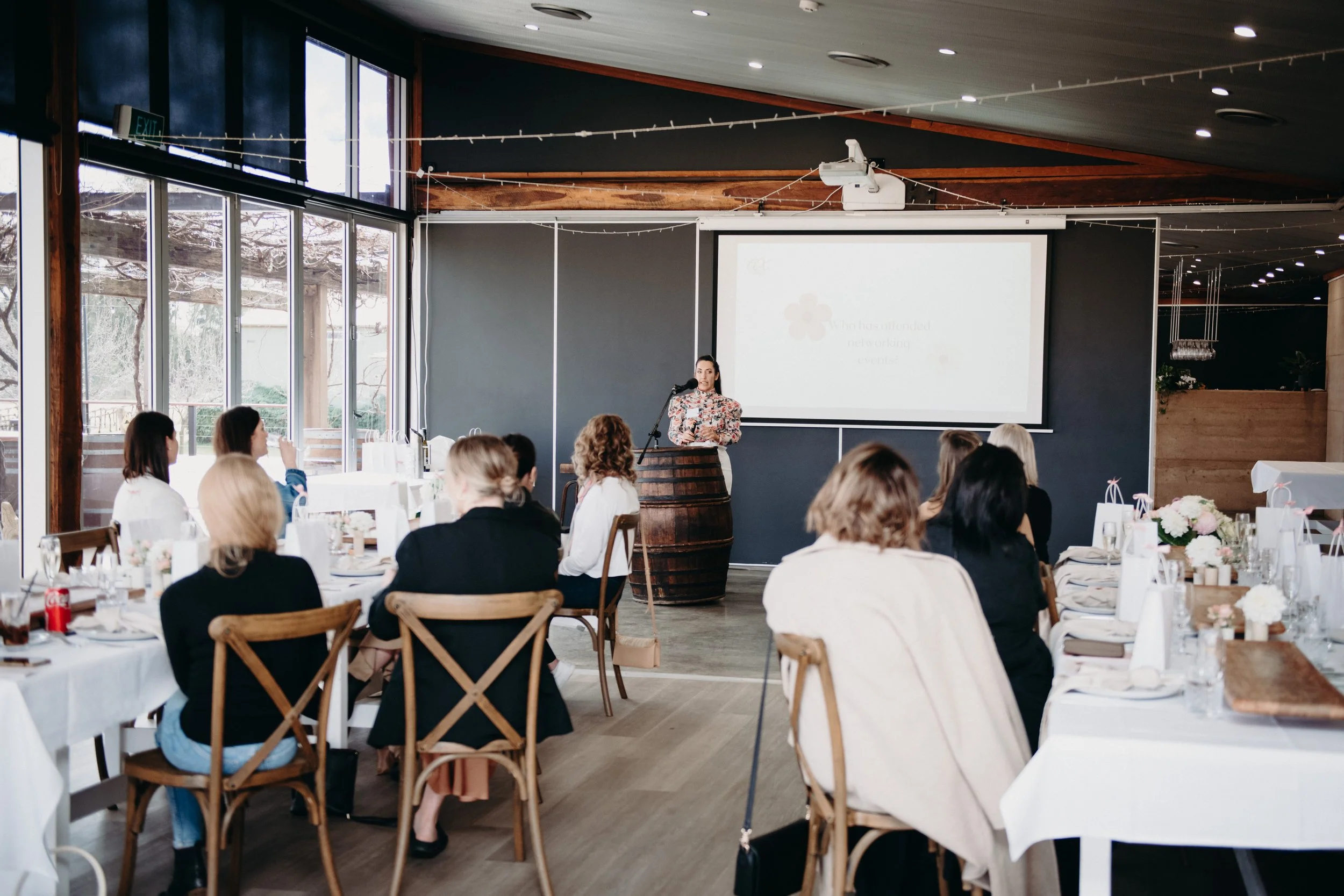 A woman gives a presentation to a group of women seated at round tables in a well-lit, modern event space with large windows.