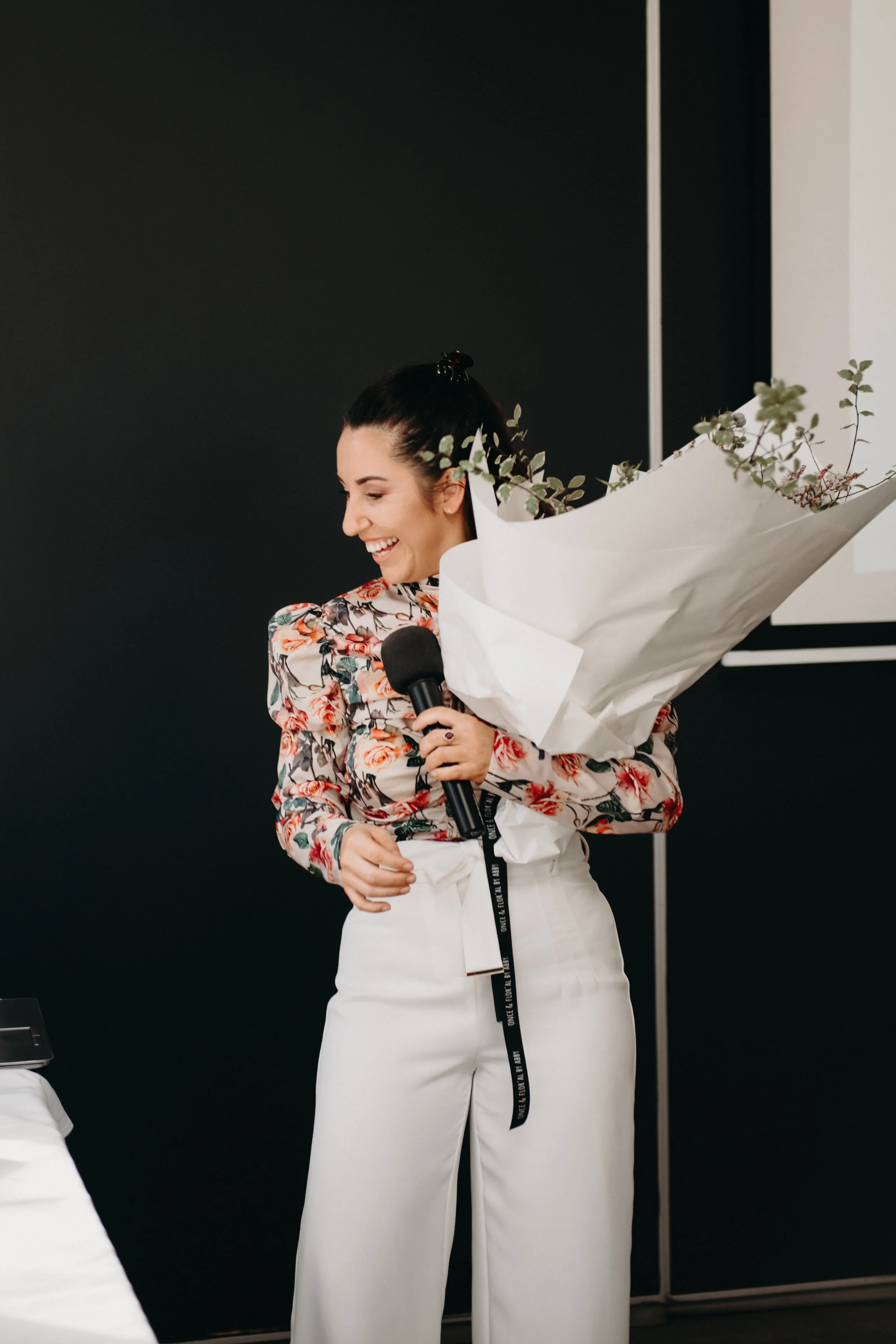 A woman holding a microphone and smiling while holding a large bouquet of flowers.