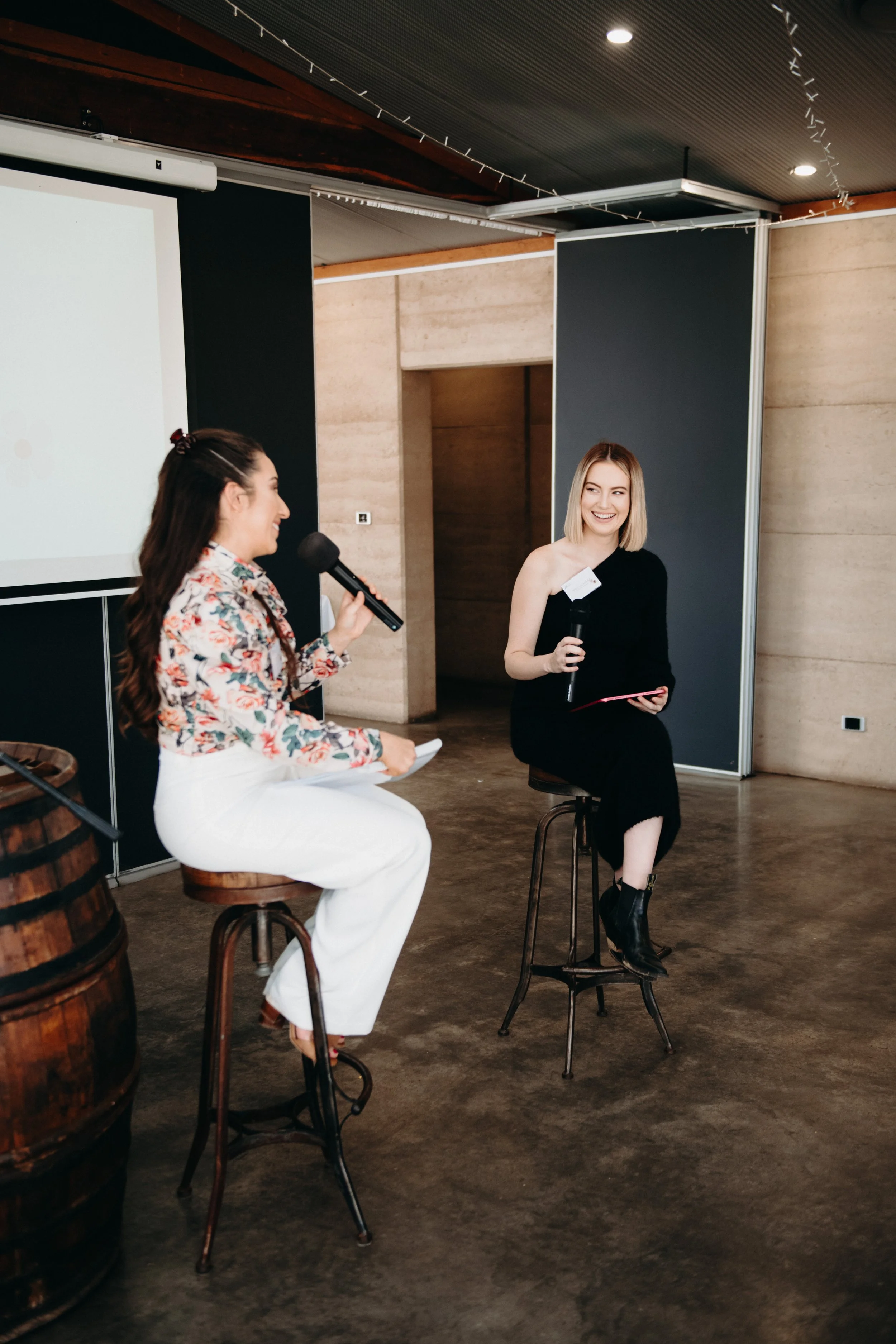 Two women in a discussion, seated on bar stools, with microphones; one in a floral blouse and white pants, the other in a black dress, in a modern indoor setting.