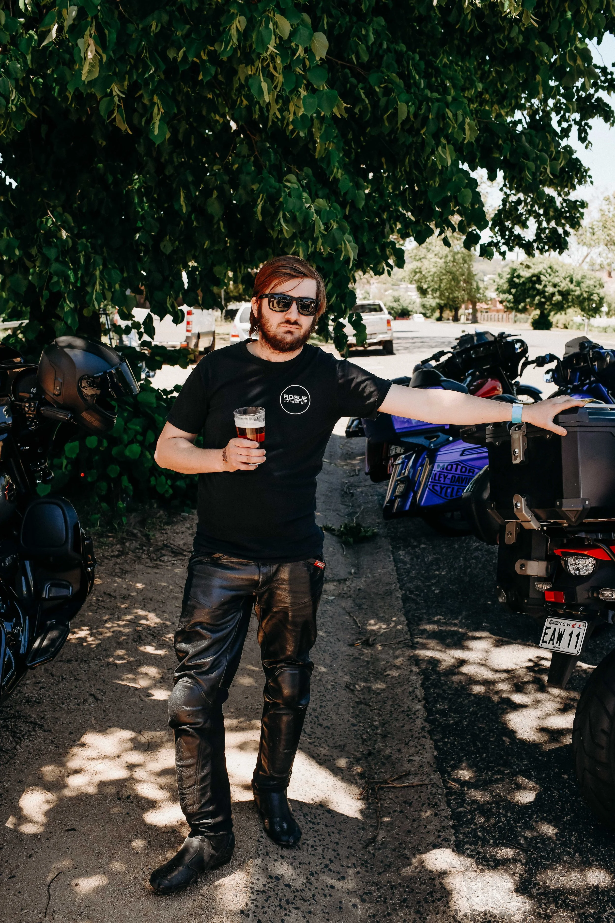 A man with shoulder-length hair, beard, and sunglasses stands outdoors under a large leafy tree, holding a glass of dark beer in his right hand. He is wearing a black T-shirt with a logo and black leather pants. Several motorcycles are parked beside him, and a parking lot with trees and vehicles is visible in the background.
