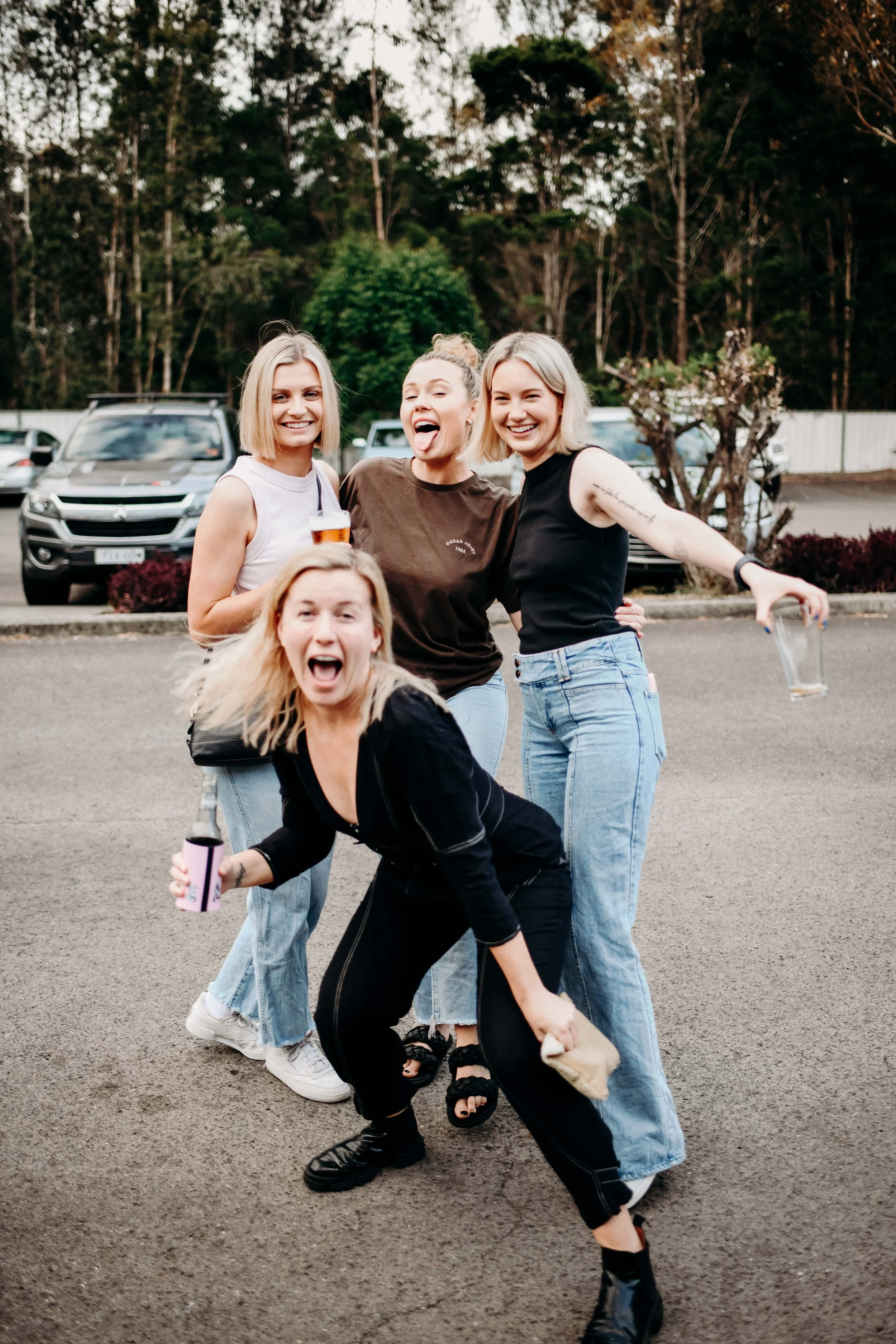 Four women smiling and cheering outdoors in a parking lot with trees in the background, holding drinks.
