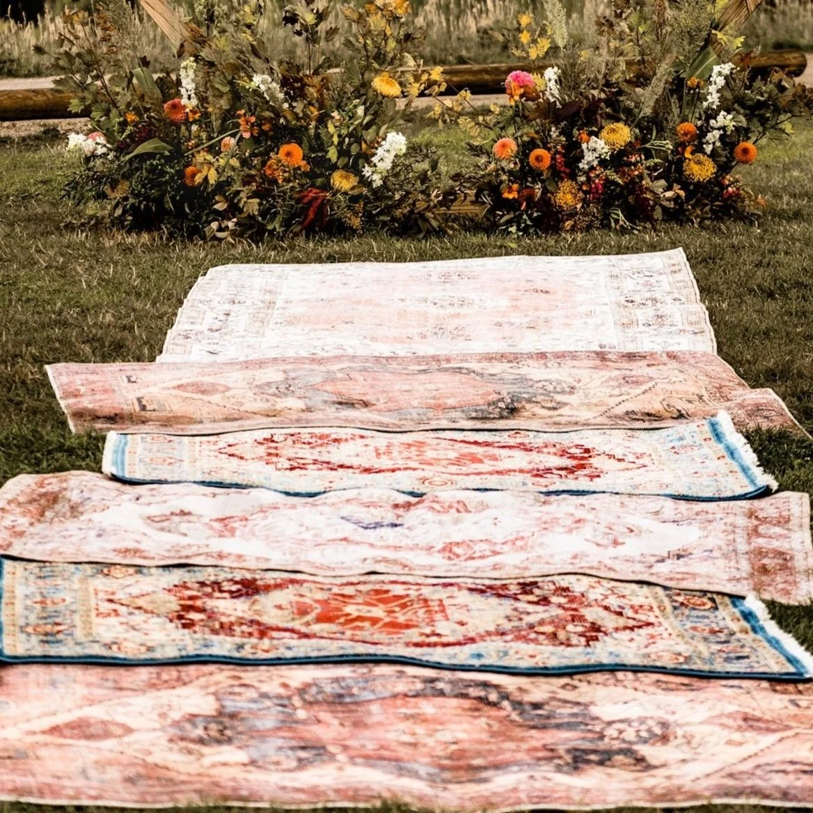 Multiple stacked vintage rugs laid out on grass with a colorful floral arrangement in the background.