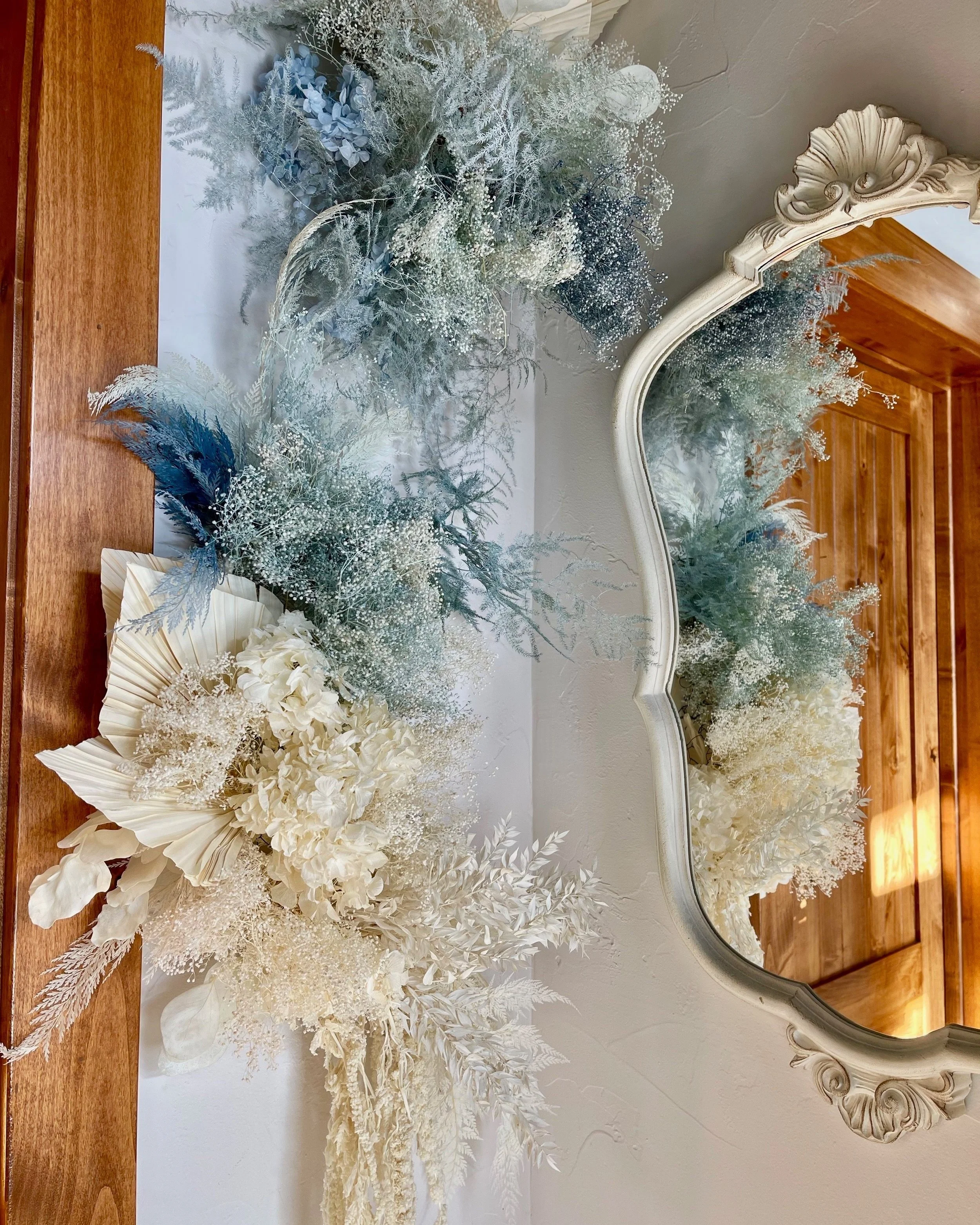 A floral arrangement of preserved flowers and foliage in white, cream, blue, and silver hues, reflected in an ornate mirror on a wooden surface.