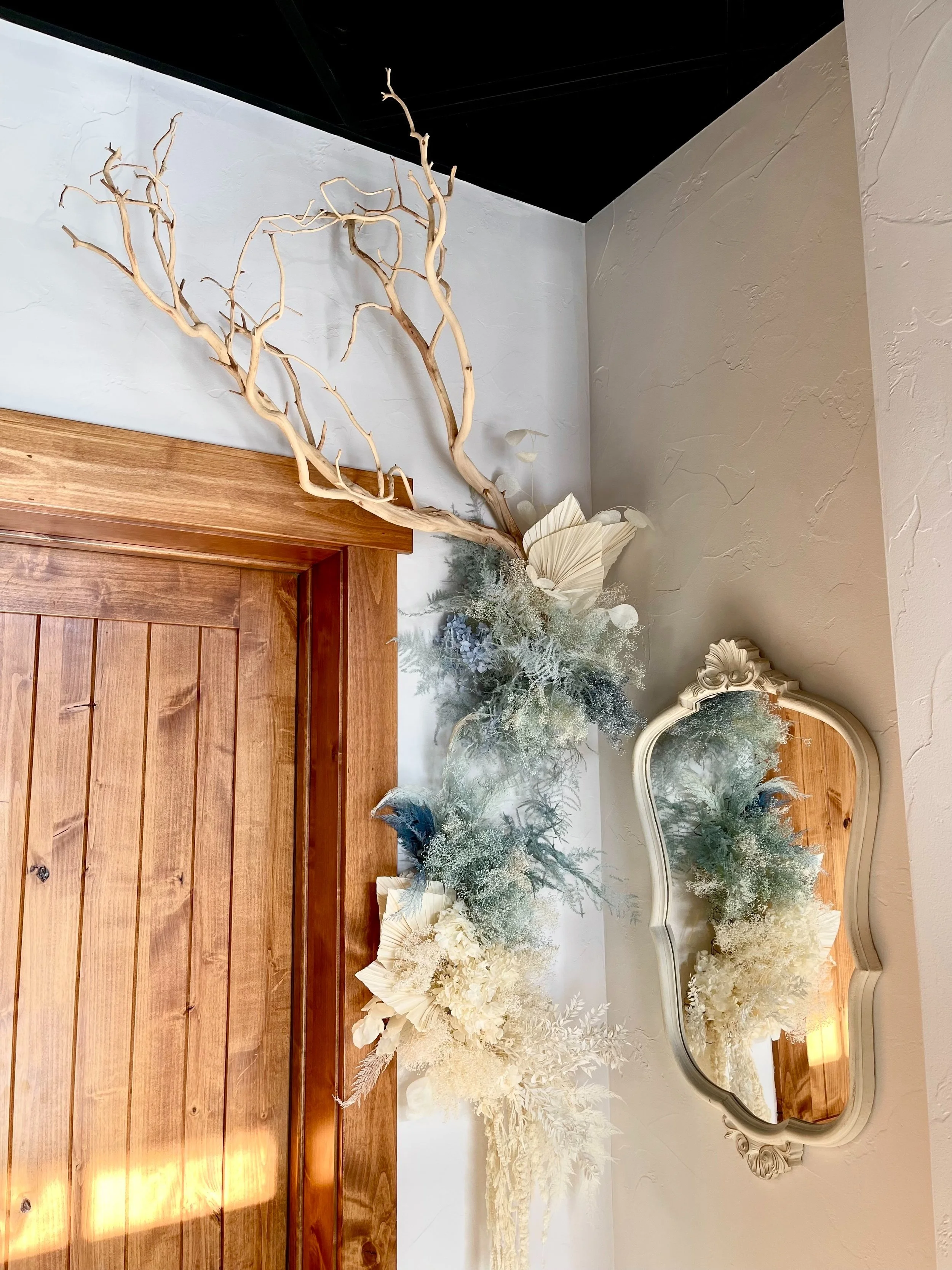 Decorative floral arrangement with dried branches, white flowers, and foliage, reflected in an ornate mirror on a white wall with a wooden door below.